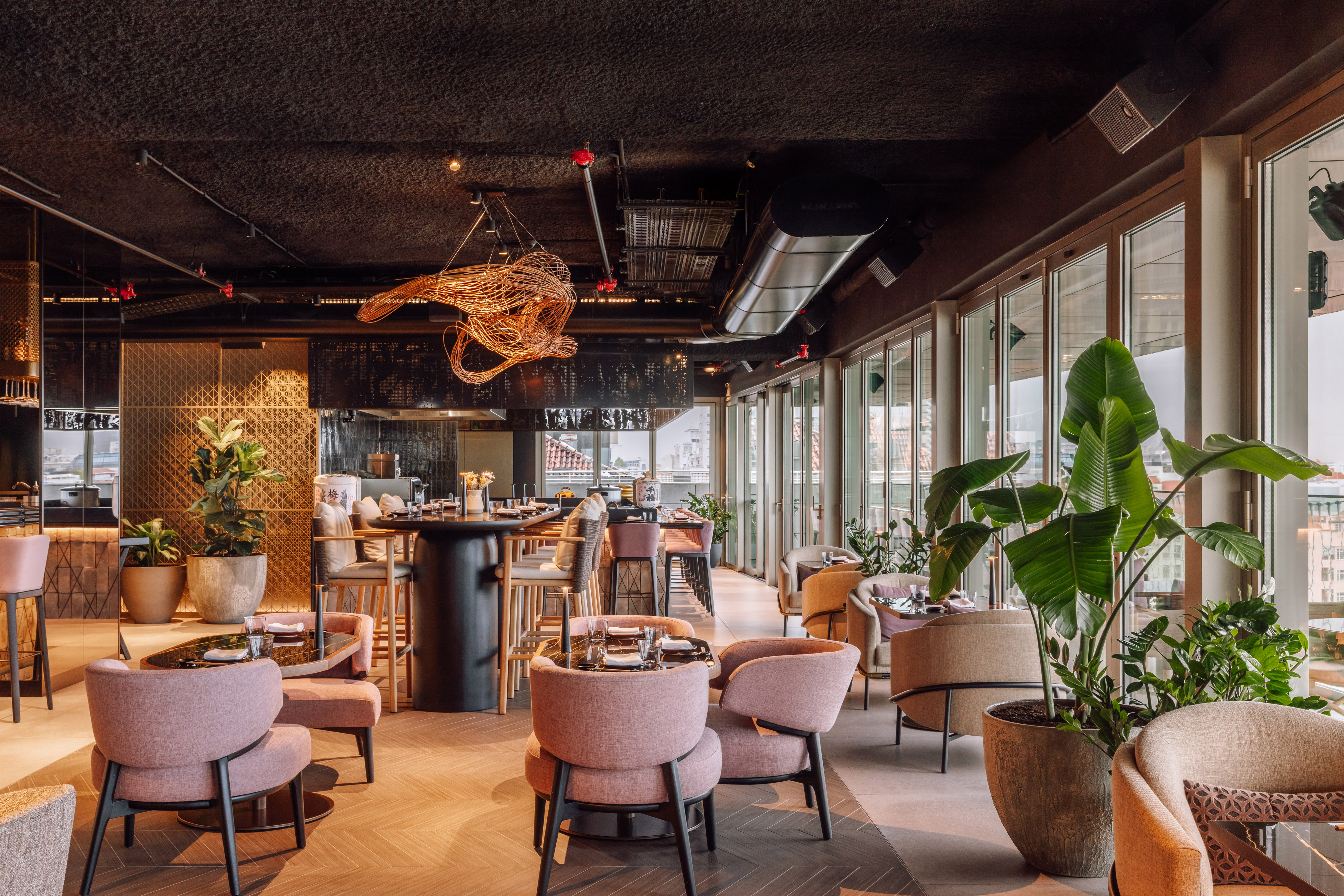 Chic modern restaurant with pink armchairs, bar, potted plants, city views.