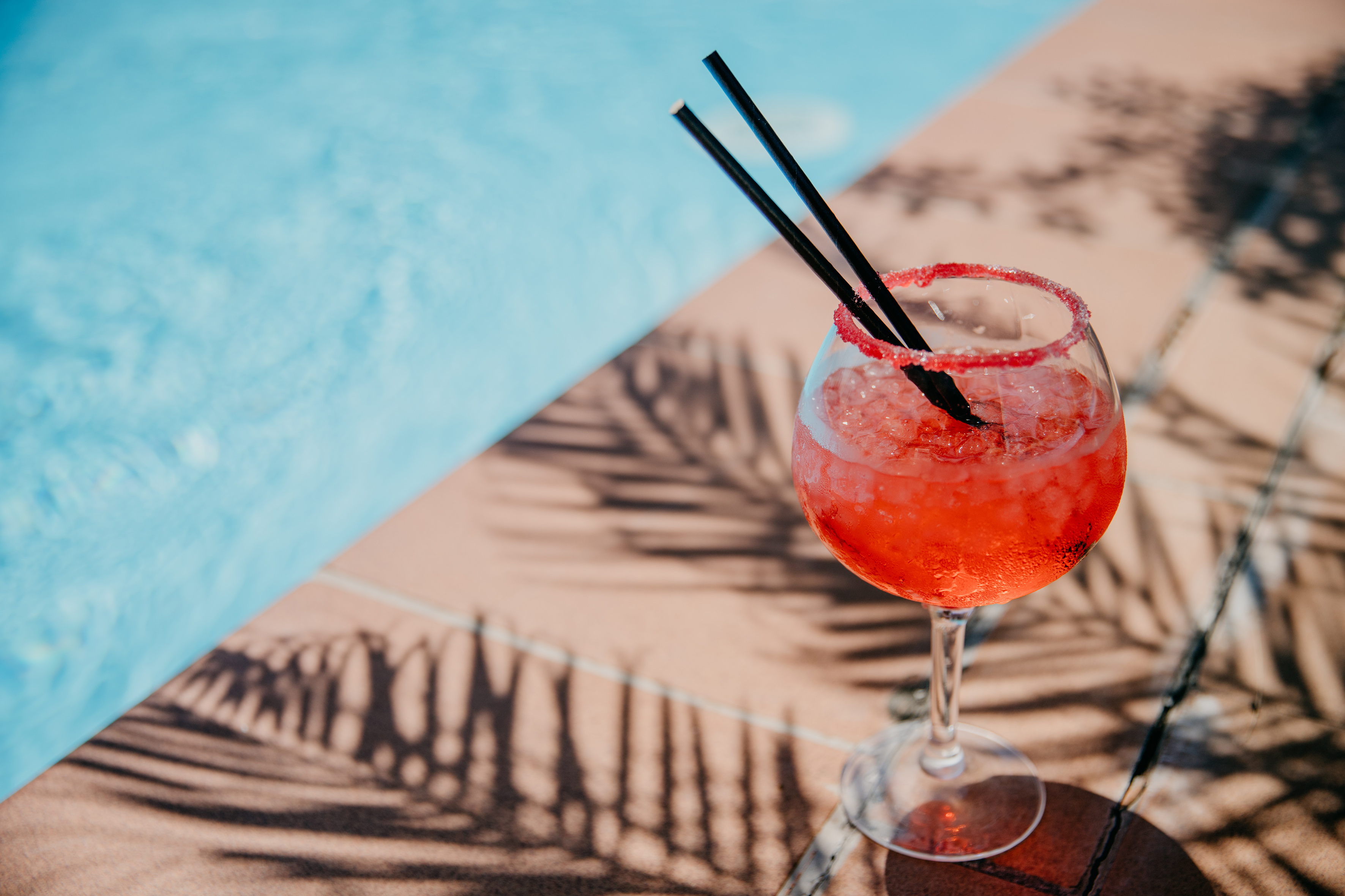 Red cocktail with sugared rim and straws, poolside with palm shadows.