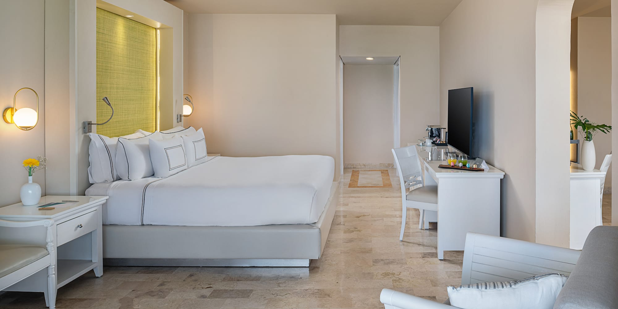 Modern hotel suite with king bed, white linens, bright decor, and stone floor.