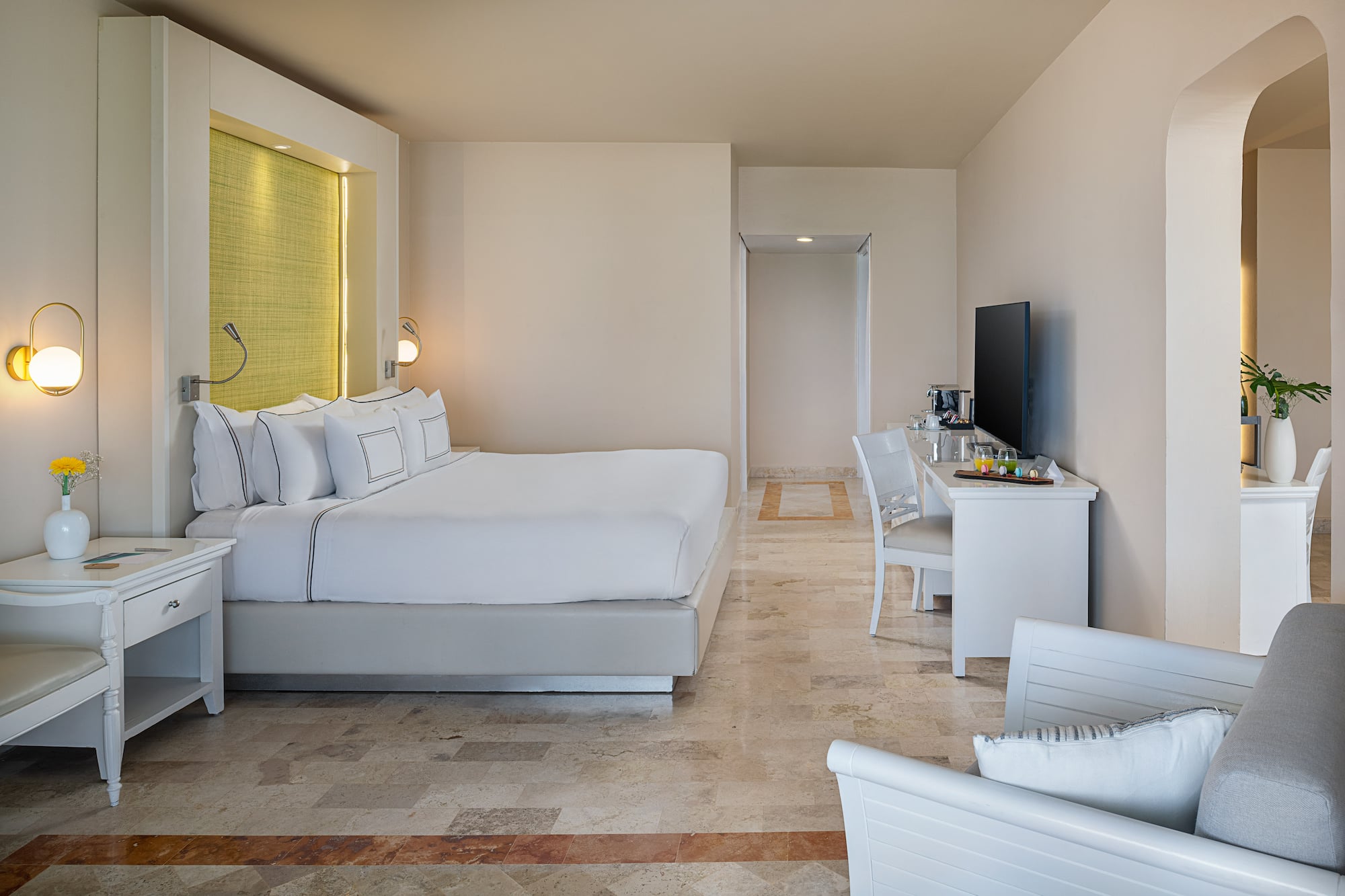 Modern hotel suite with king bed, white linens, bright decor, and stone floor.