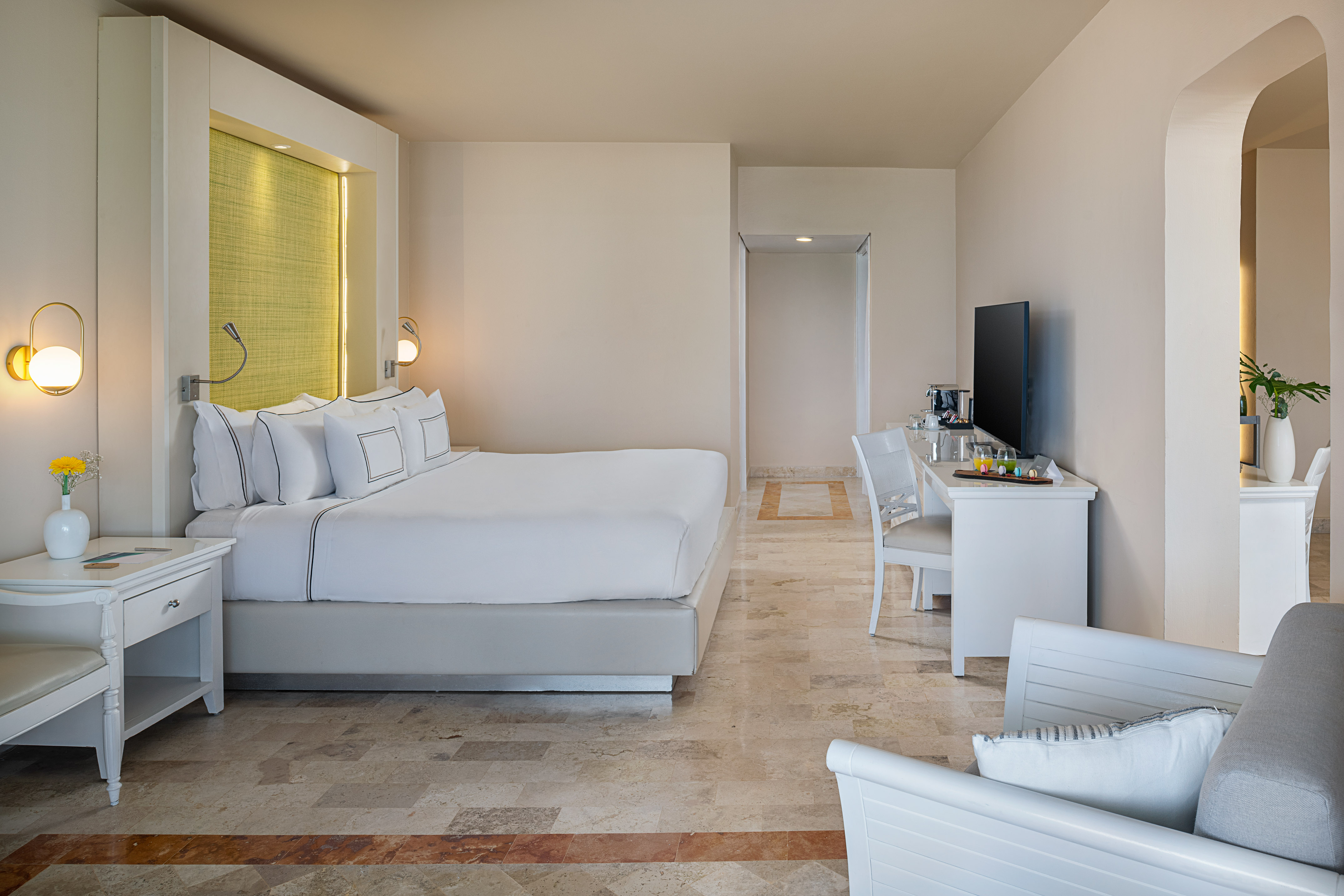 Modern hotel suite with king bed, white linens, bright decor, and stone floor.
