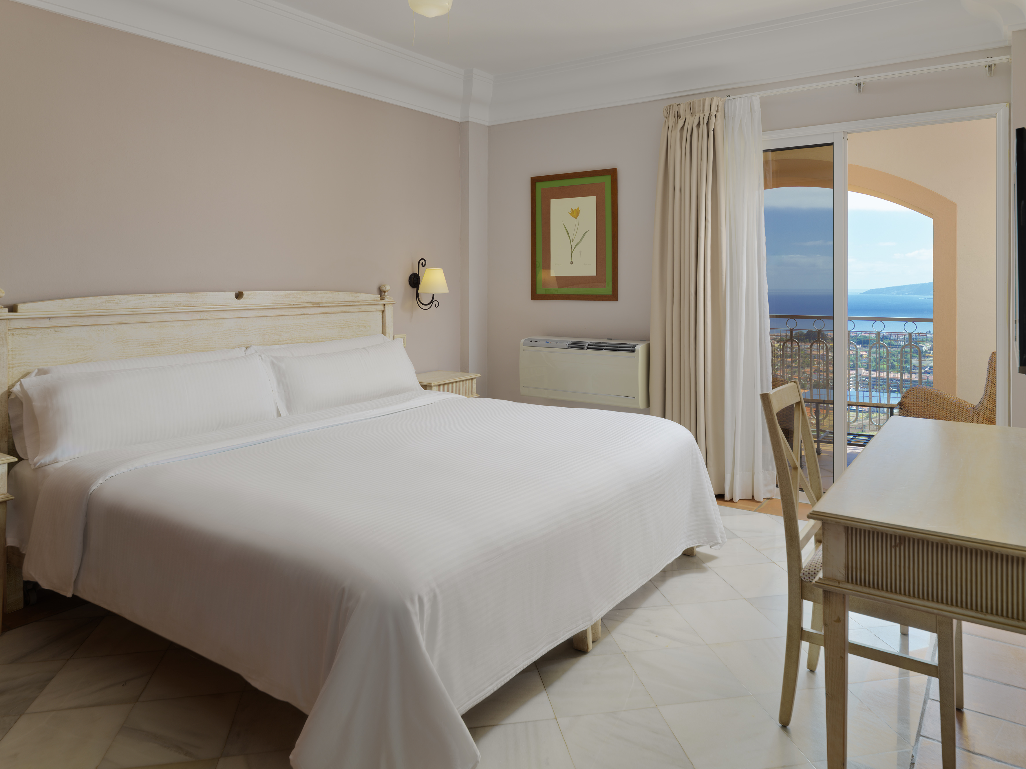Hotel bedroom with white bed, sea view balcony, desk, and tiled floor.