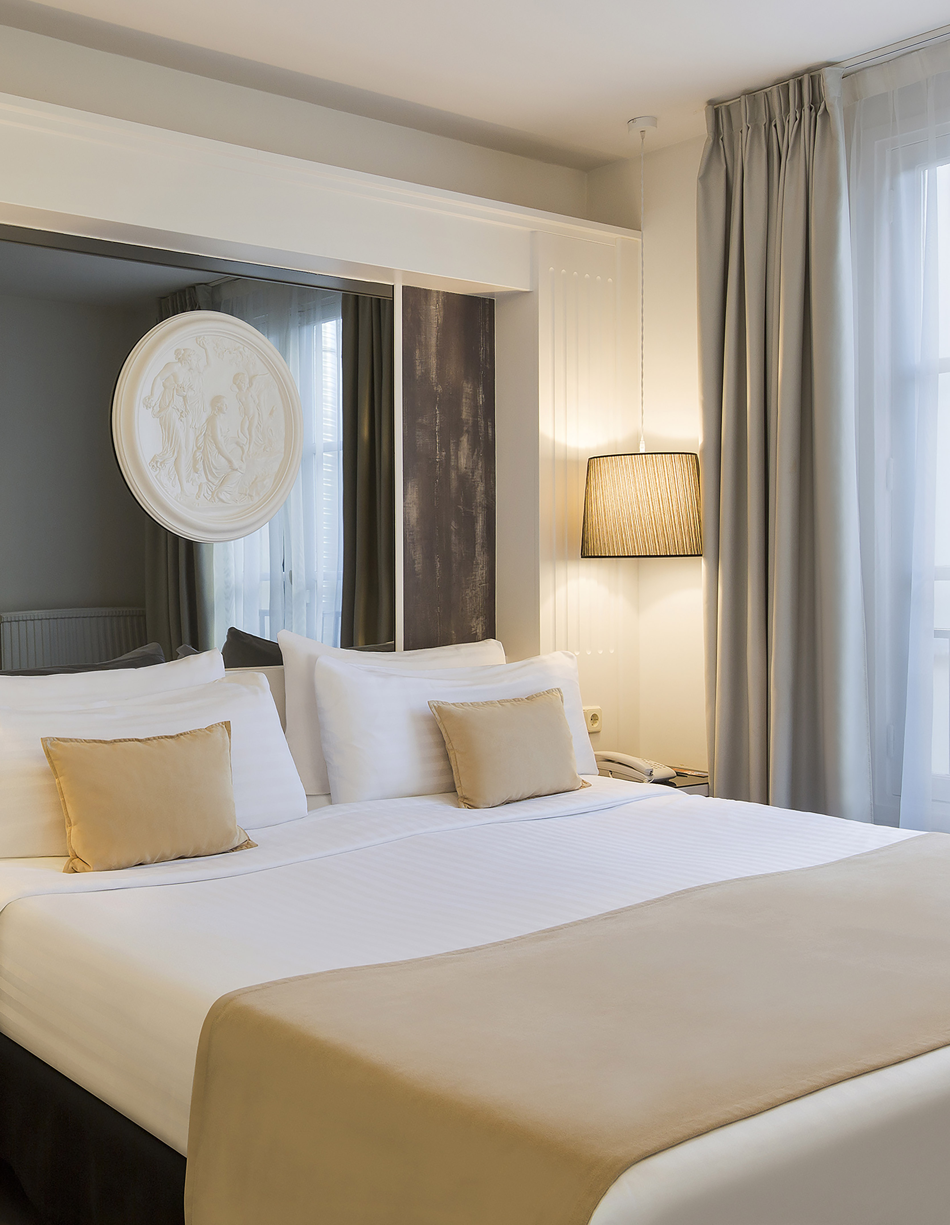 Modern hotel bedroom with white linens, gold pillows, hanging lamp, and window.