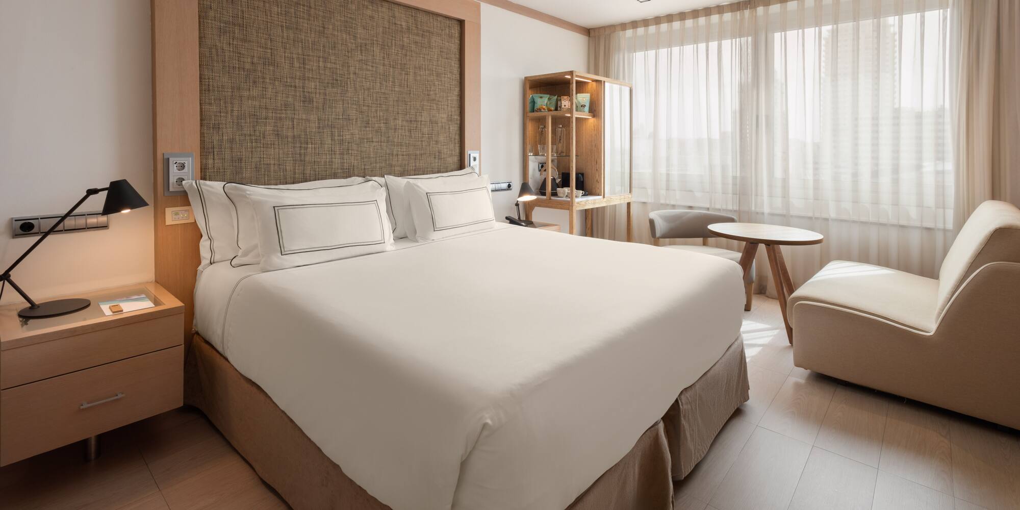 Modern hotel room with bed, wood accents, nightstand, and bright window.