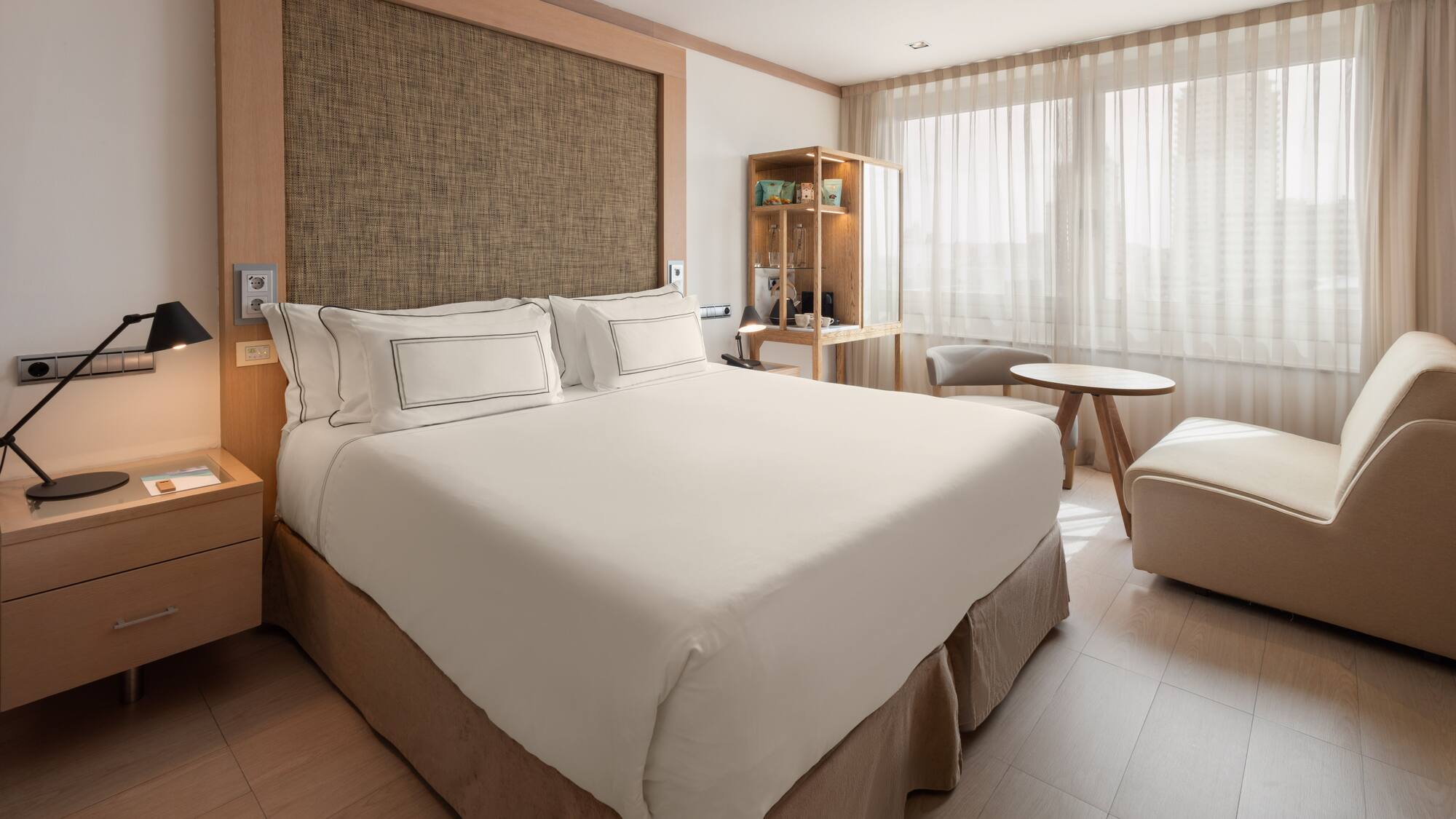 Modern hotel room with bed, wood accents, nightstand, and bright window.