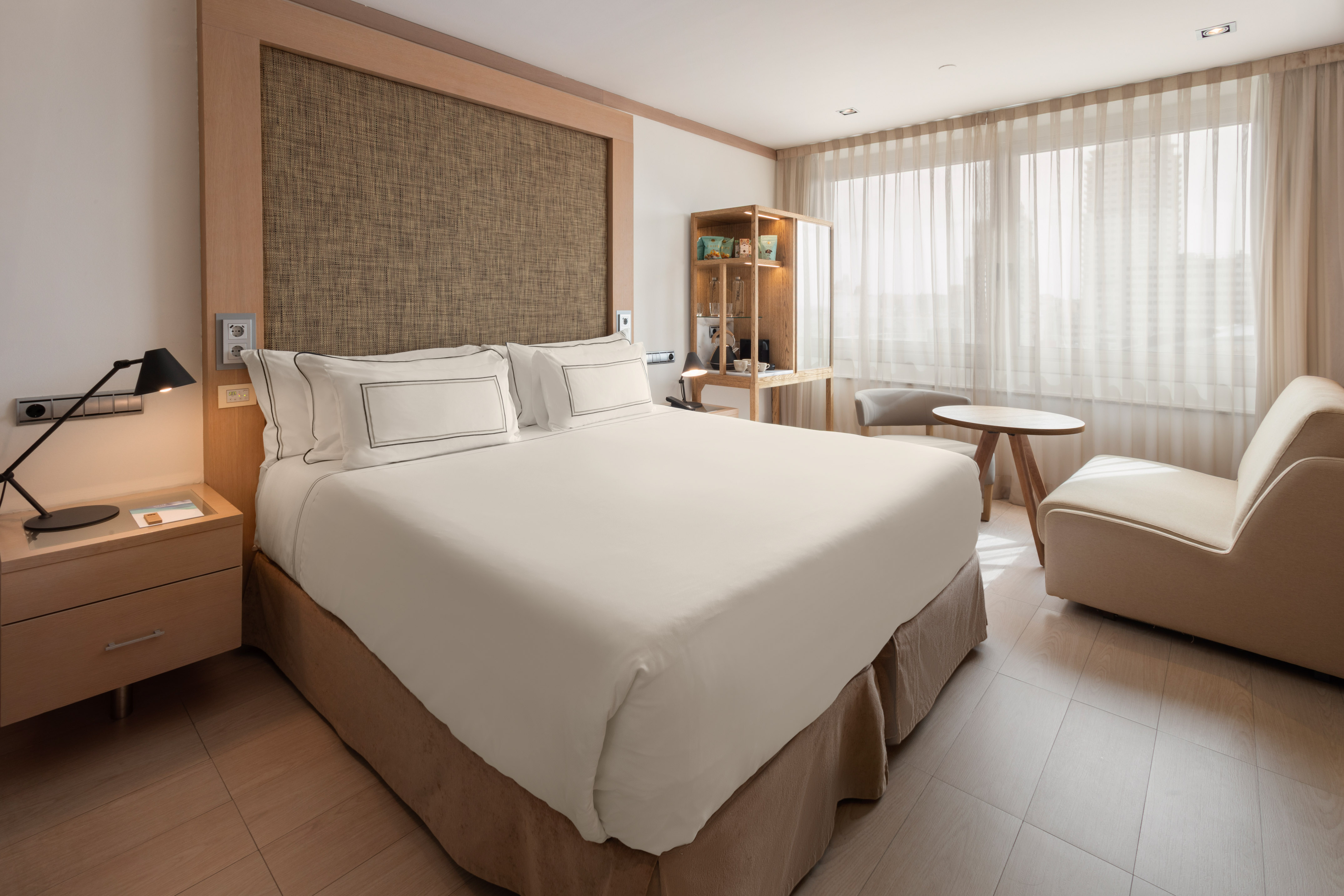 Modern hotel room with bed, wood accents, nightstand, and bright window.