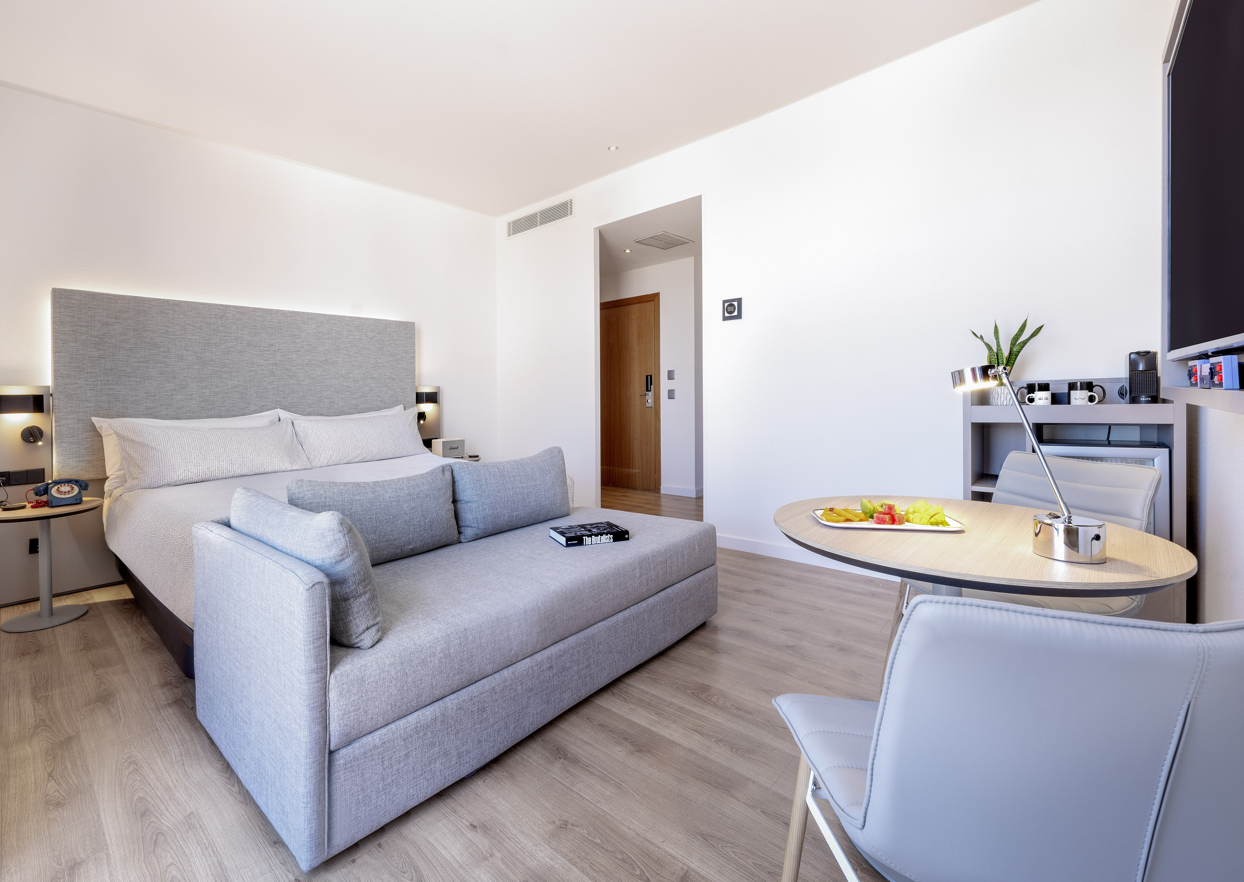 Bright hotel studio with bed, grey sofa, dining table, and wood floor.