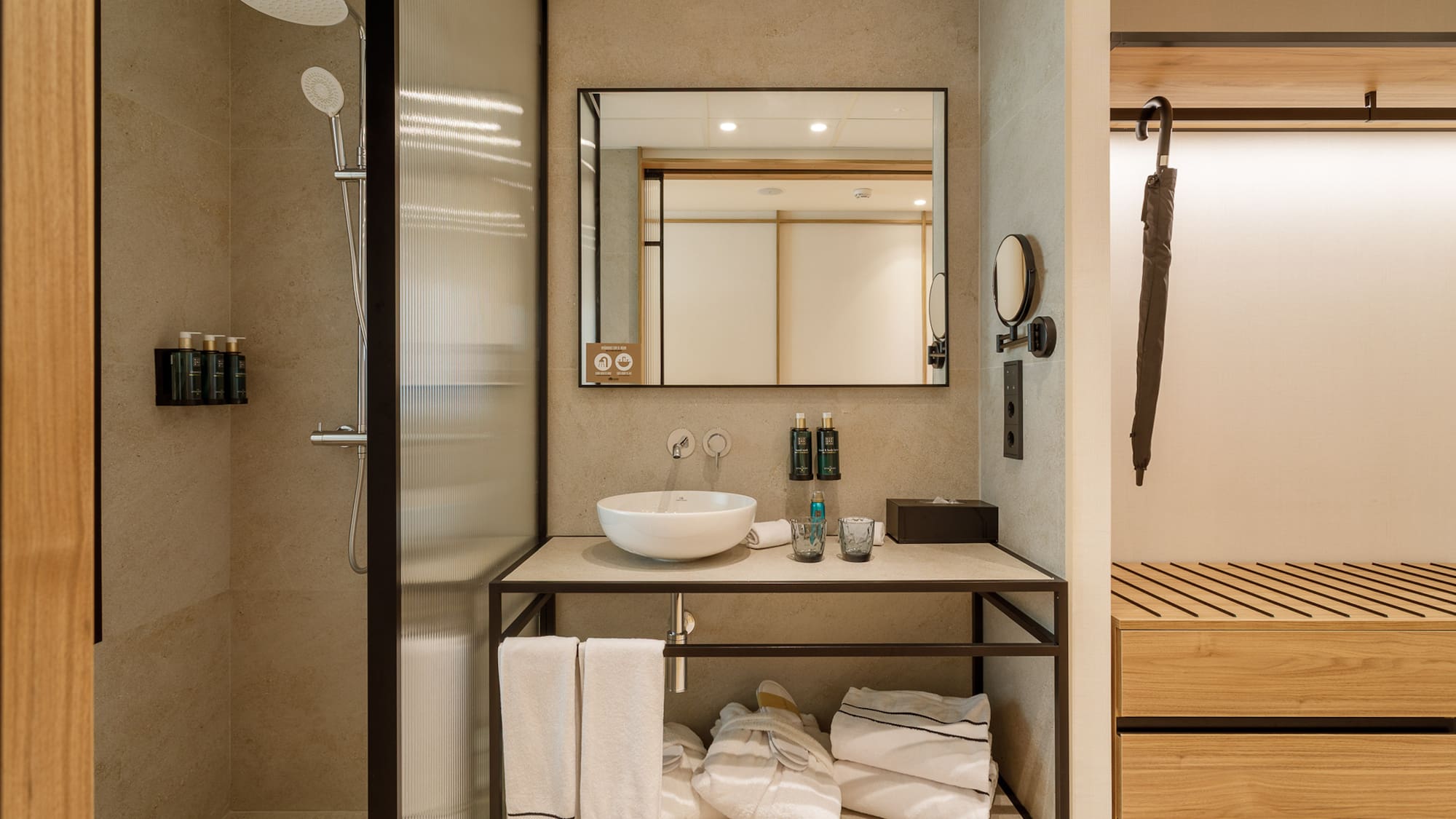 Neutral-toned hotel bathroom with glass shower, modern vanity, and closet storage.