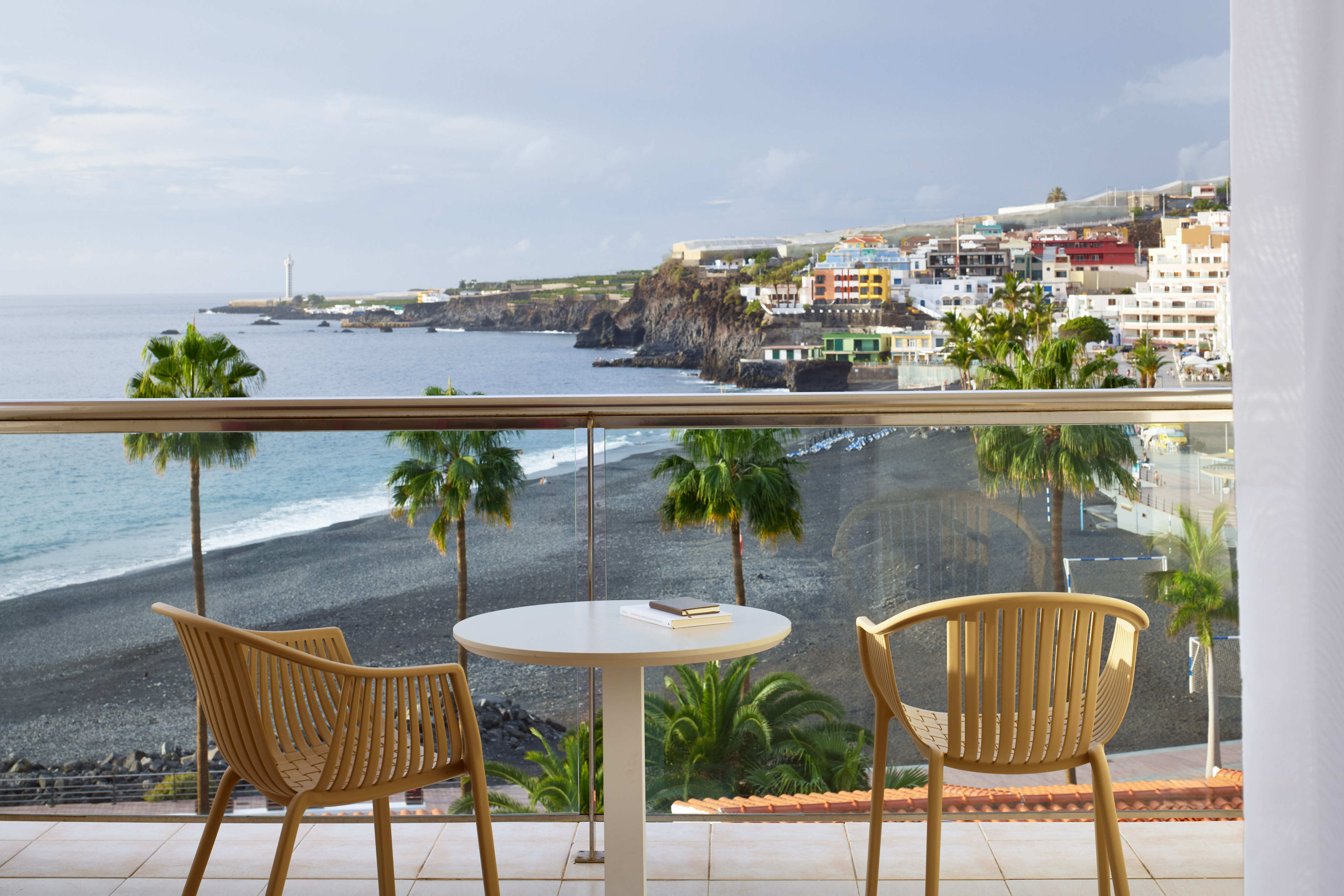 Balcony overlooking black sand beach, ocean, palm trees, and colorful coastal town.