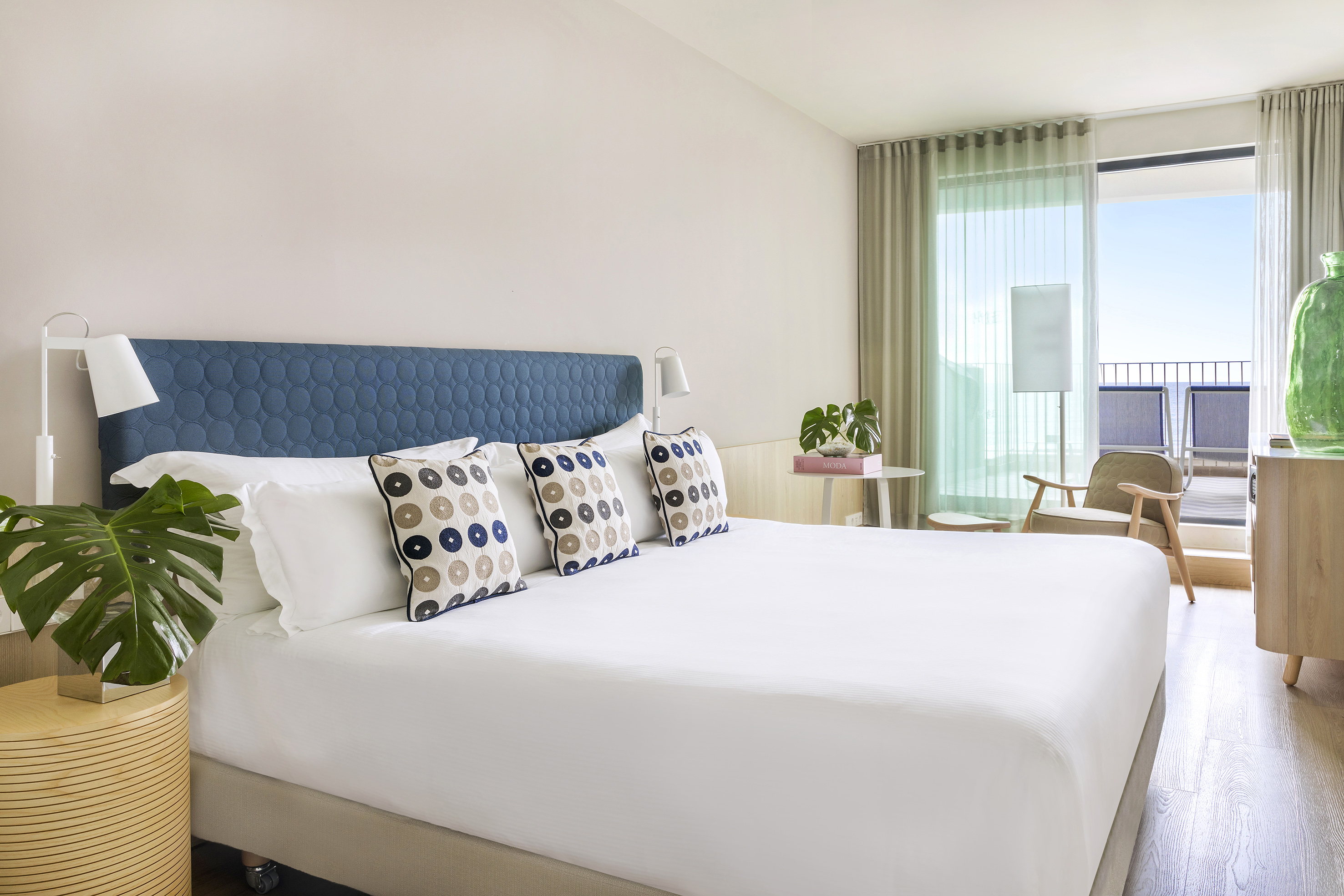 Modern hotel bedroom with white bed, blue headboard, and balcony access.