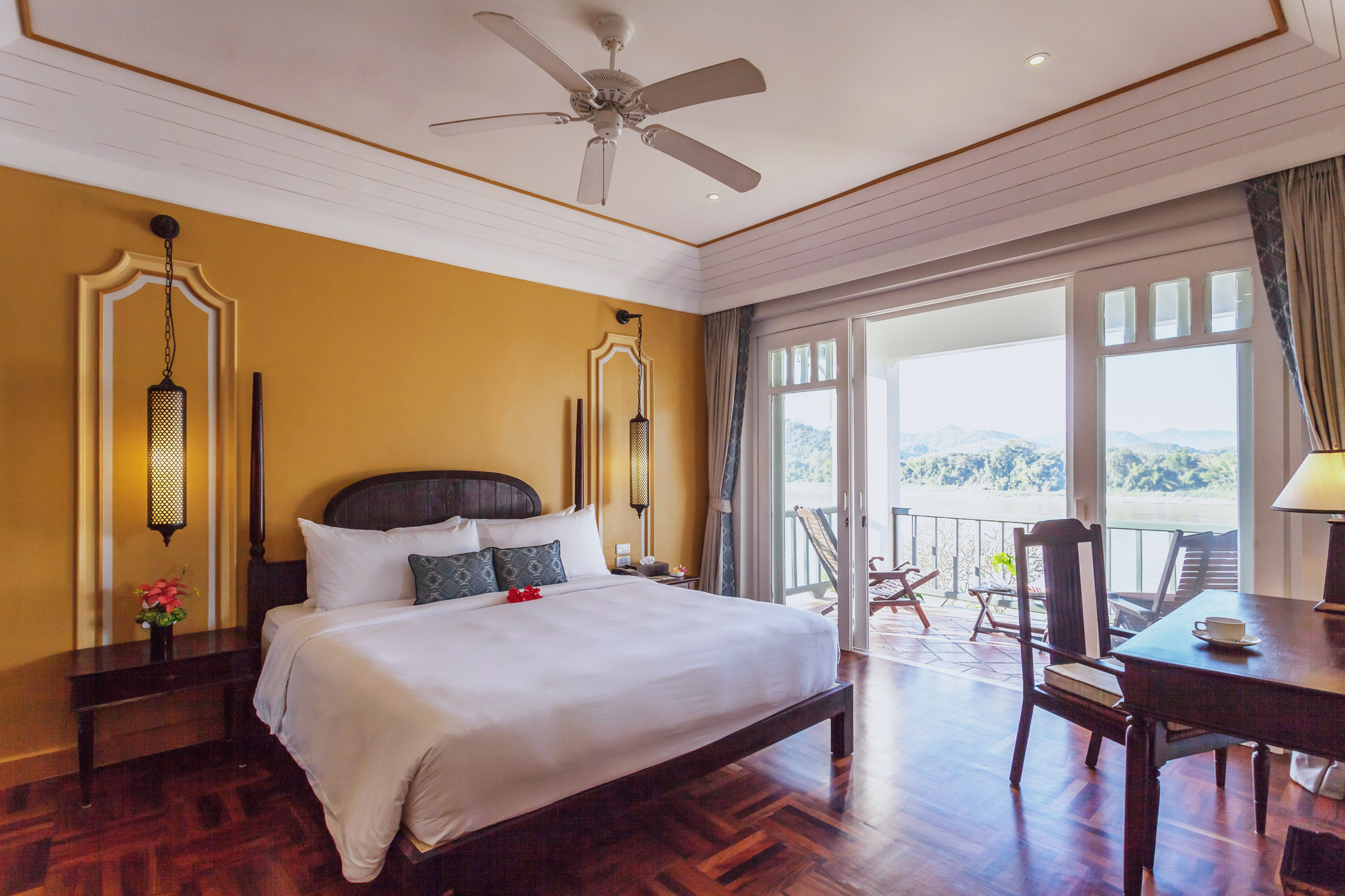 Resort bedroom with balcony overlooking river, golden walls, and dark wood.