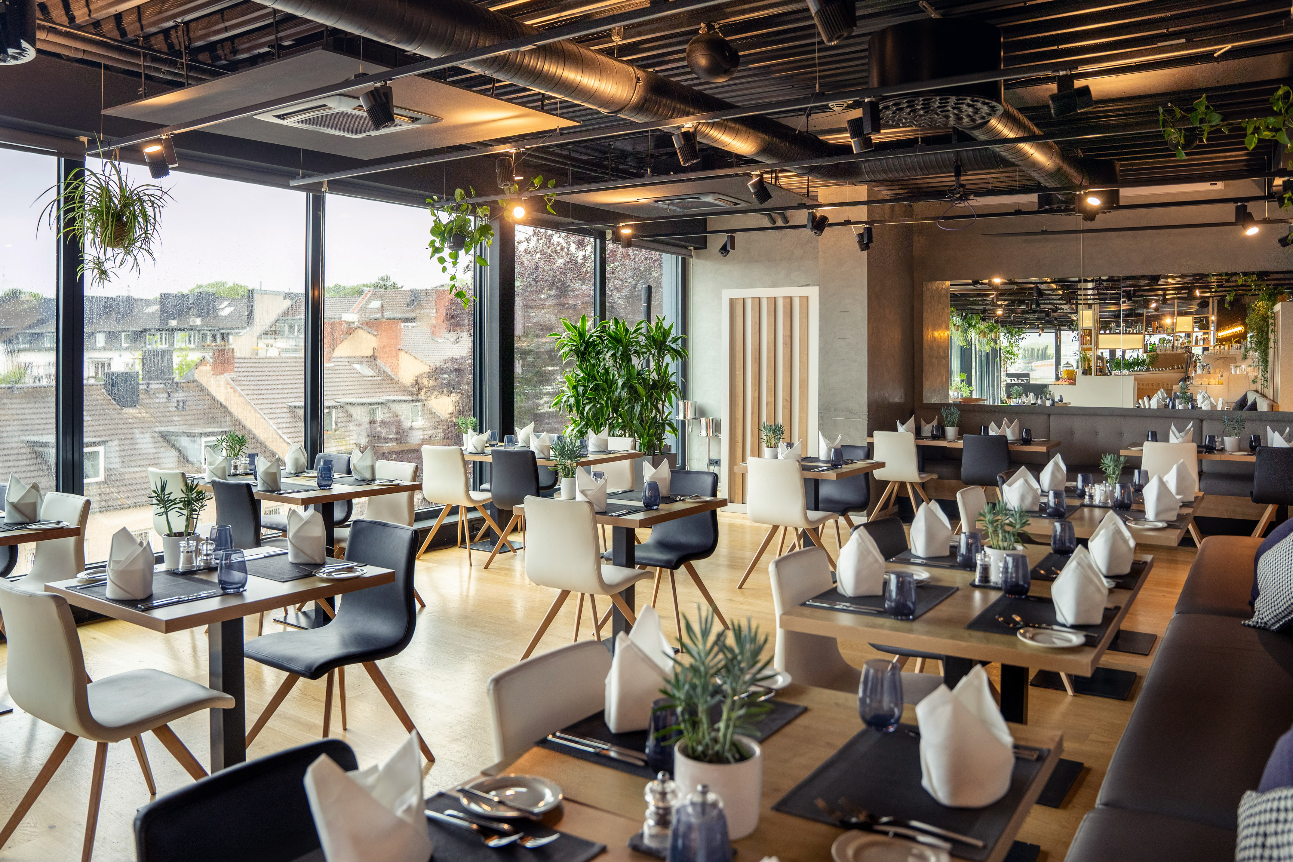Modern restaurant dining room with set tables, city view, and industrial ceiling.