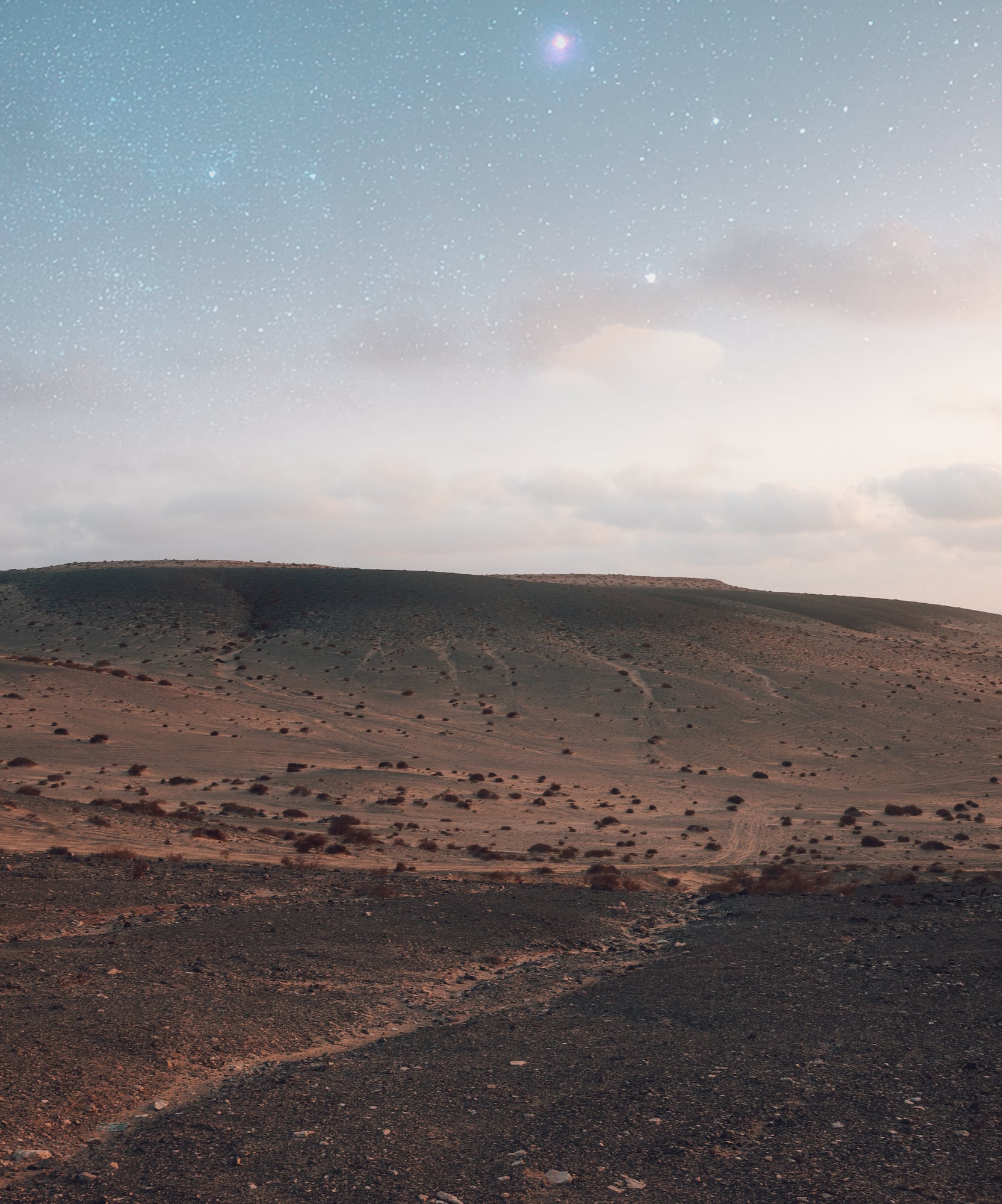 Desierto árido rocoso bajo cielo estrellado al anochecer, con mar lejano.