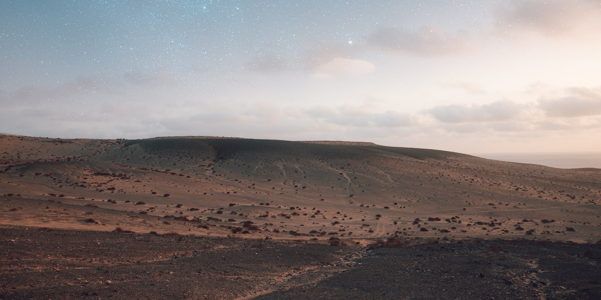 Paisaje desértico árido bajo cielo nocturno estrellado, con océano lejano.