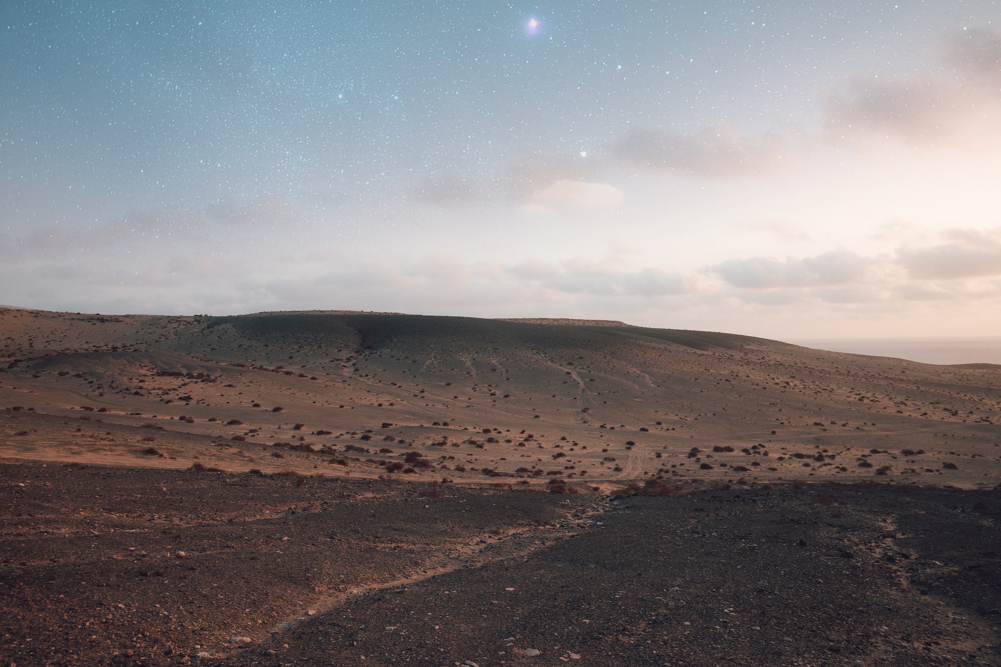 Desierto árido rocoso bajo cielo estrellado al anochecer, con mar lejano.