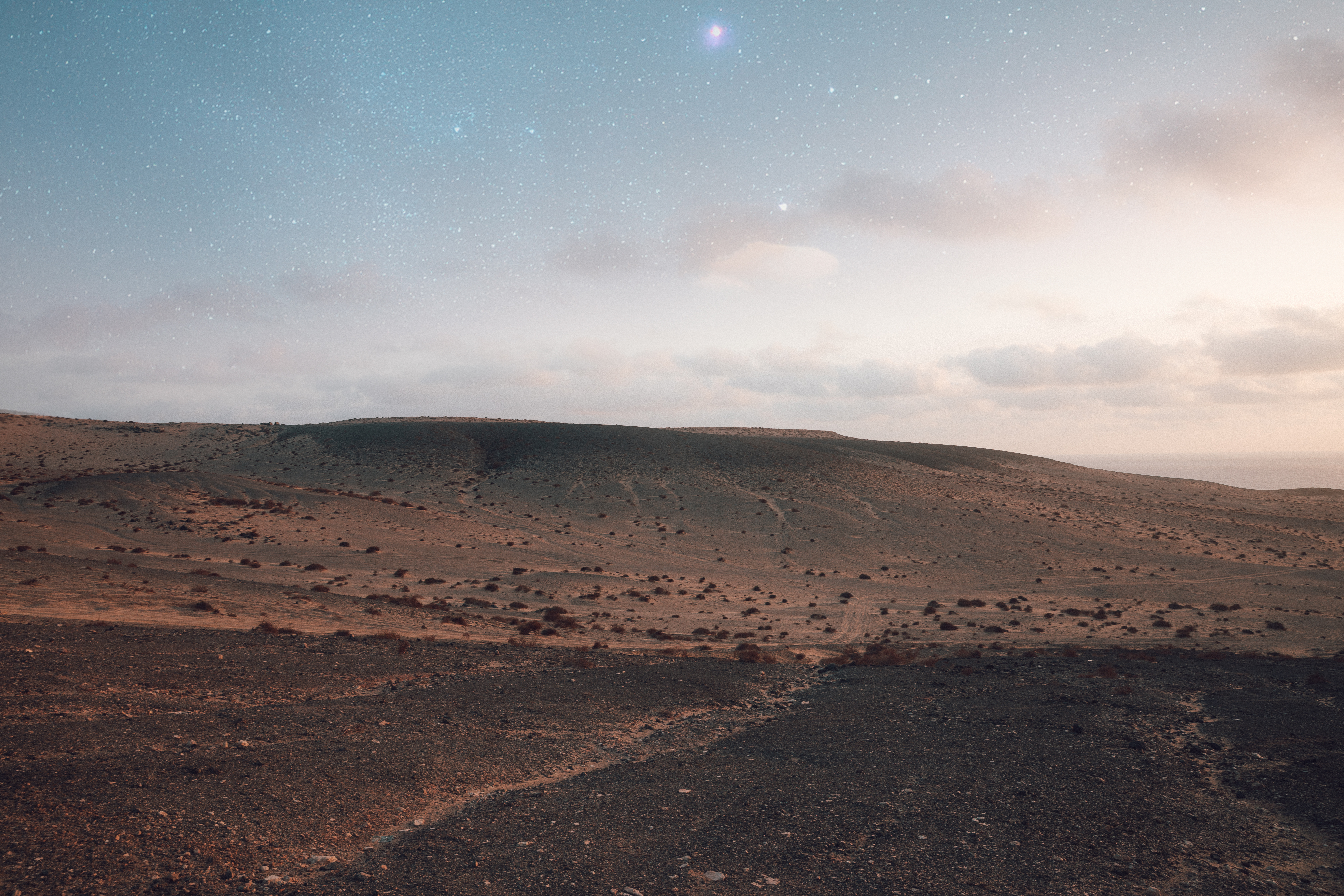 Paisaje desértico árido bajo cielo nocturno estrellado, con océano lejano.