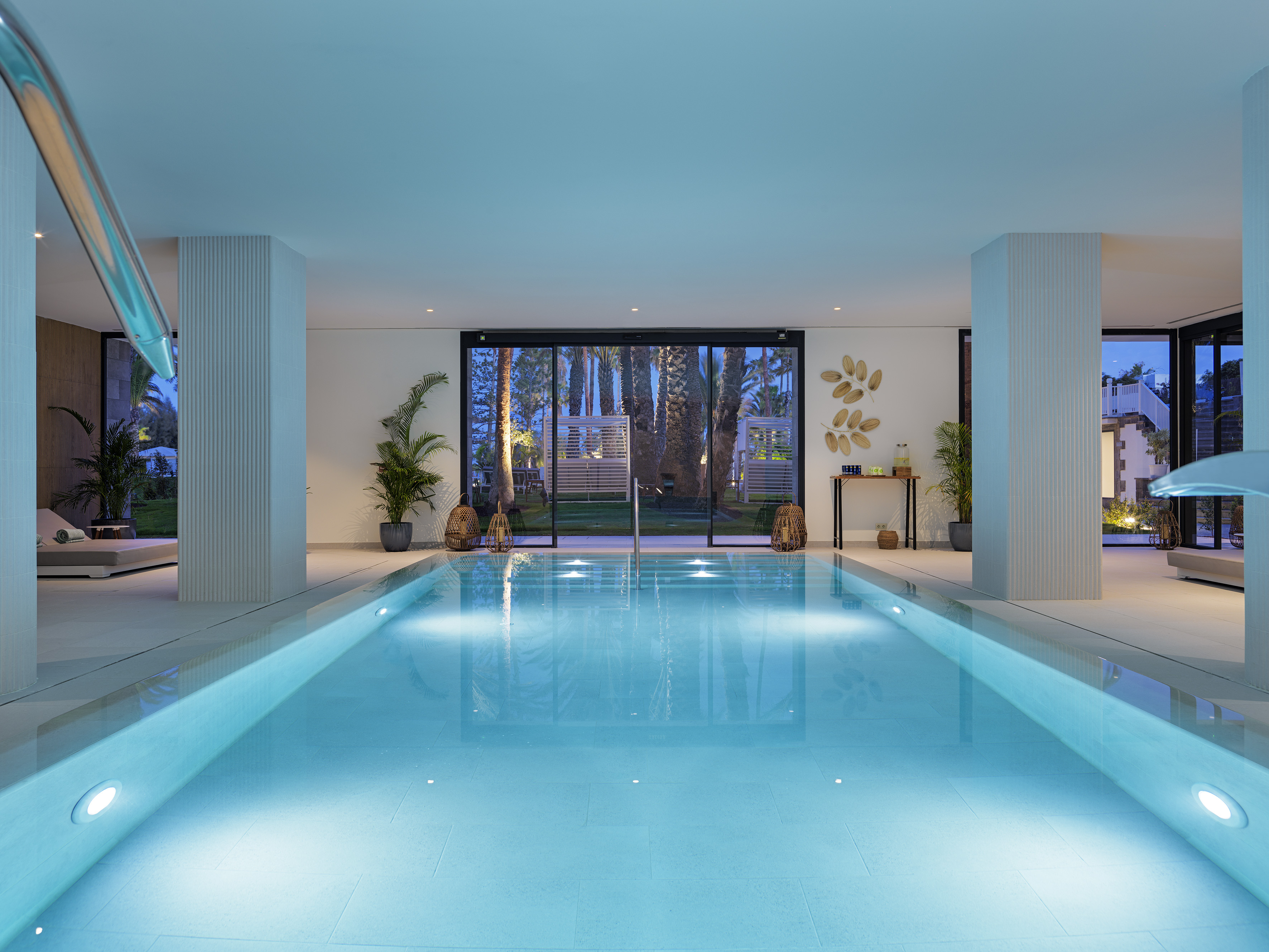 Luxurious indoor swimming pool with blue lighting and outdoor palm view.