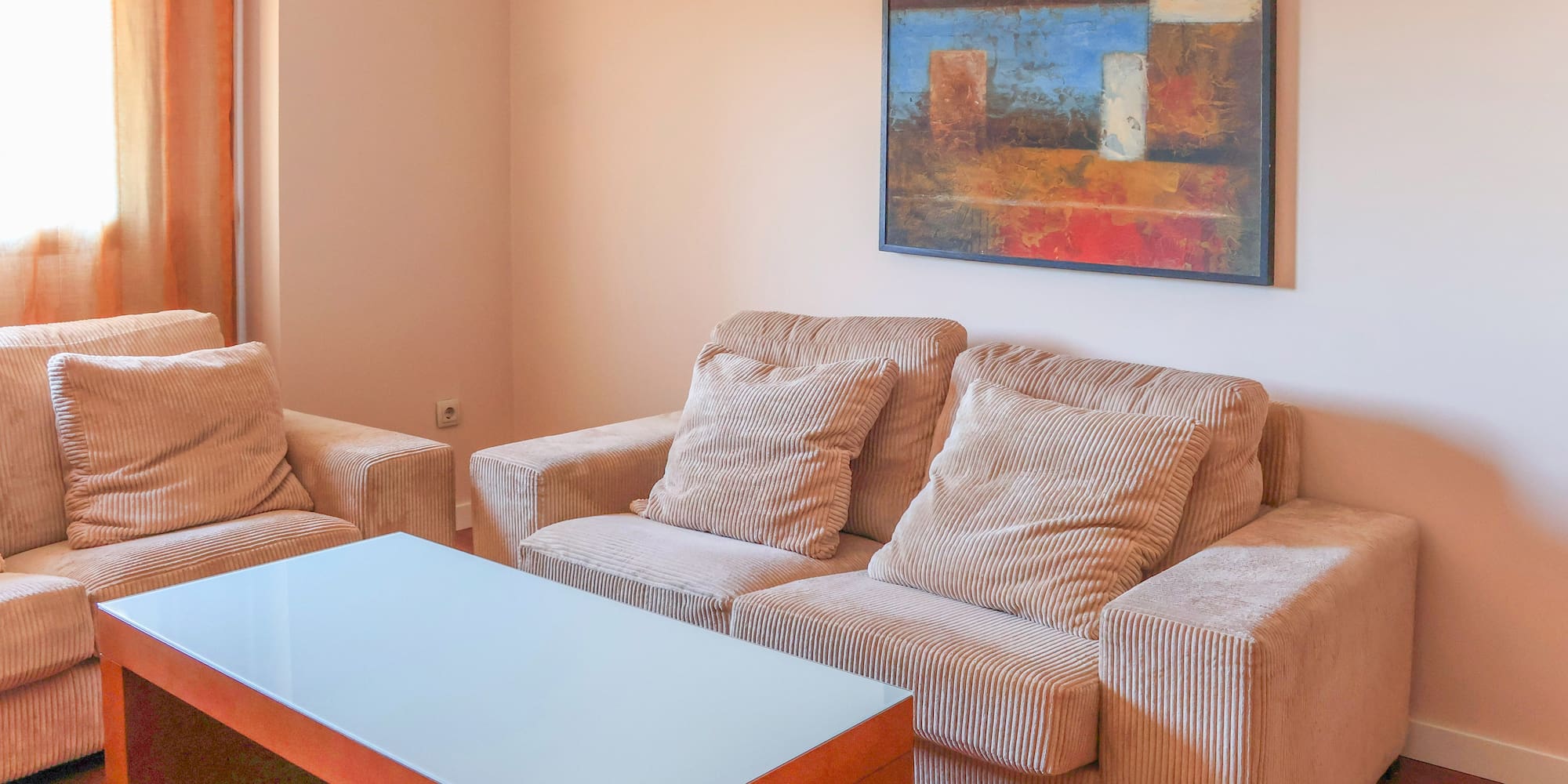 Modern living room with beige corduroy sofas, glass coffee table, and abstract art.