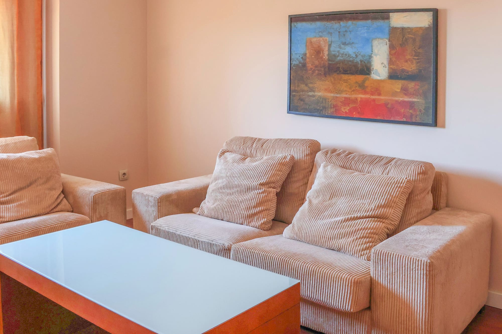 Modern living room with beige corduroy sofas, glass coffee table, and abstract art.