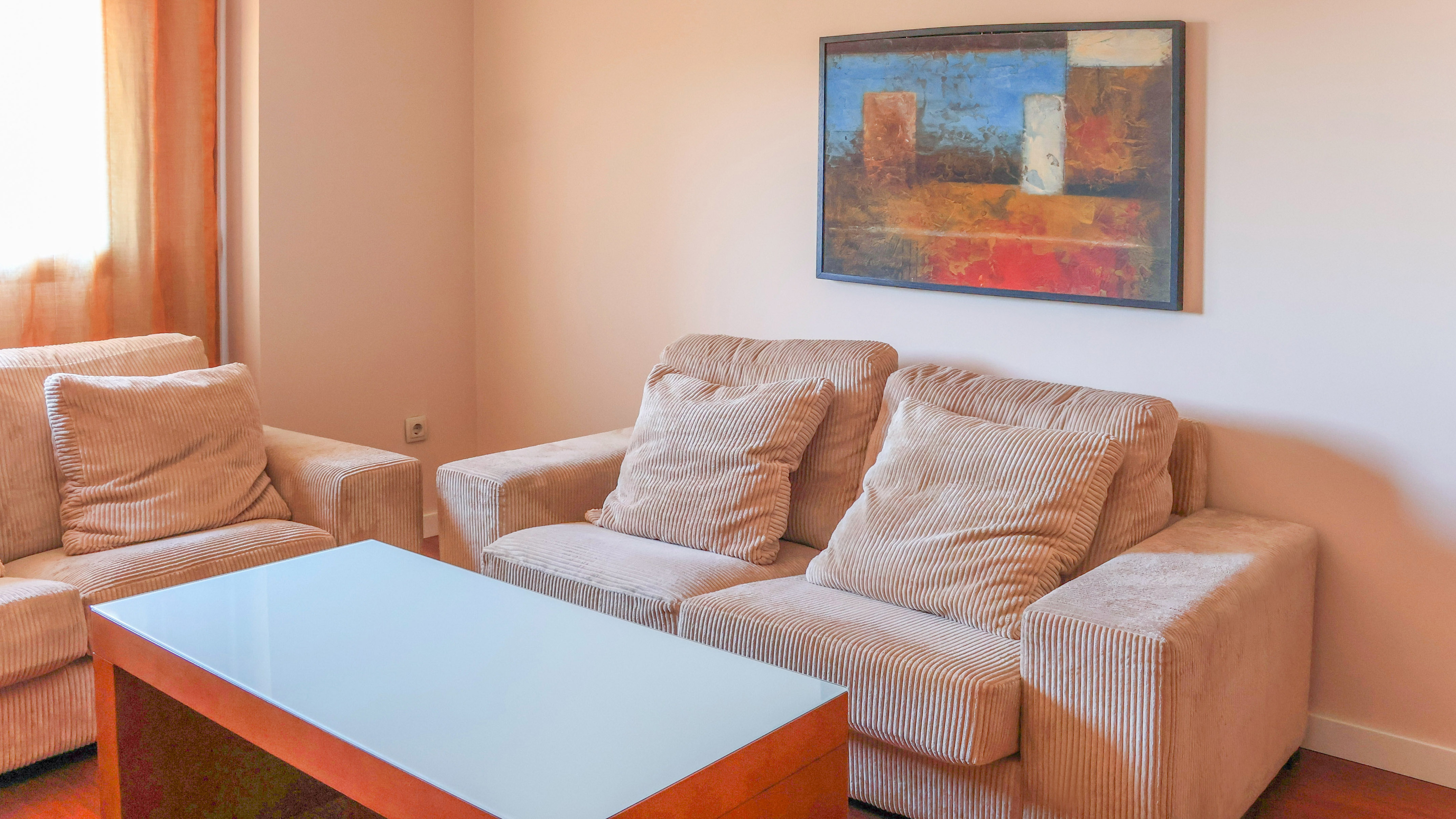 Modern living room with beige corduroy sofas, glass coffee table, and abstract art.