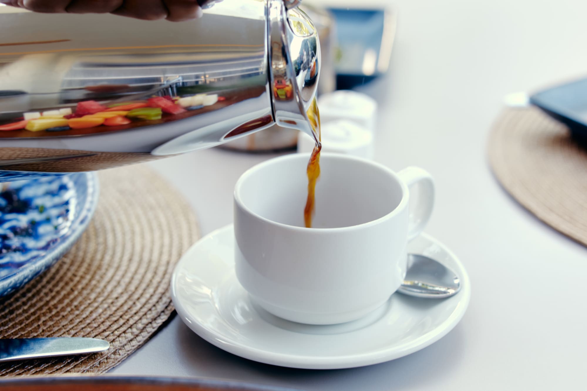 Mano vertiendo café caliente de termo a taza blanca, junto a fruta fresca.