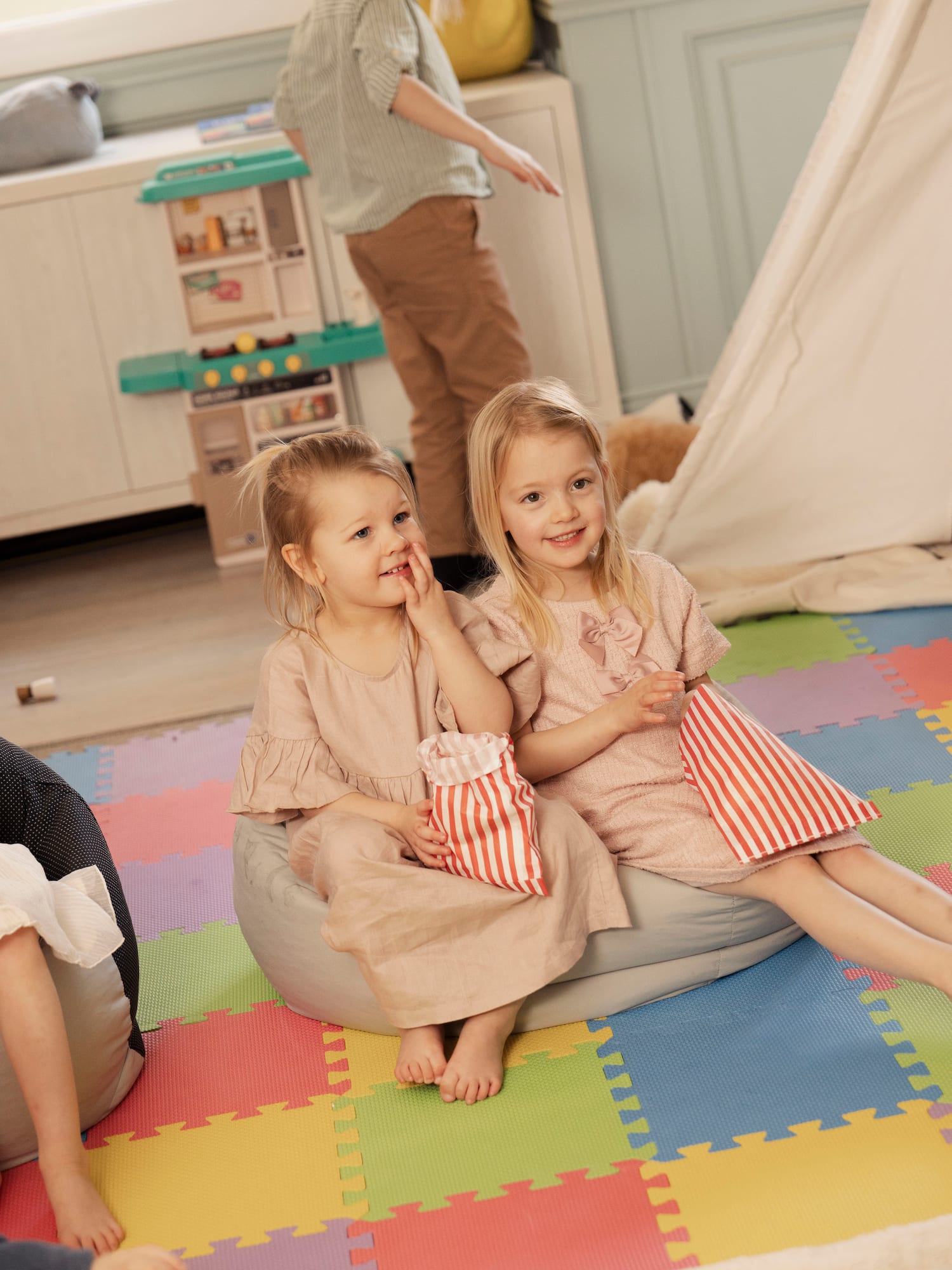 Two blonde girls on beanbags holding striped bags in a colorful playroom.