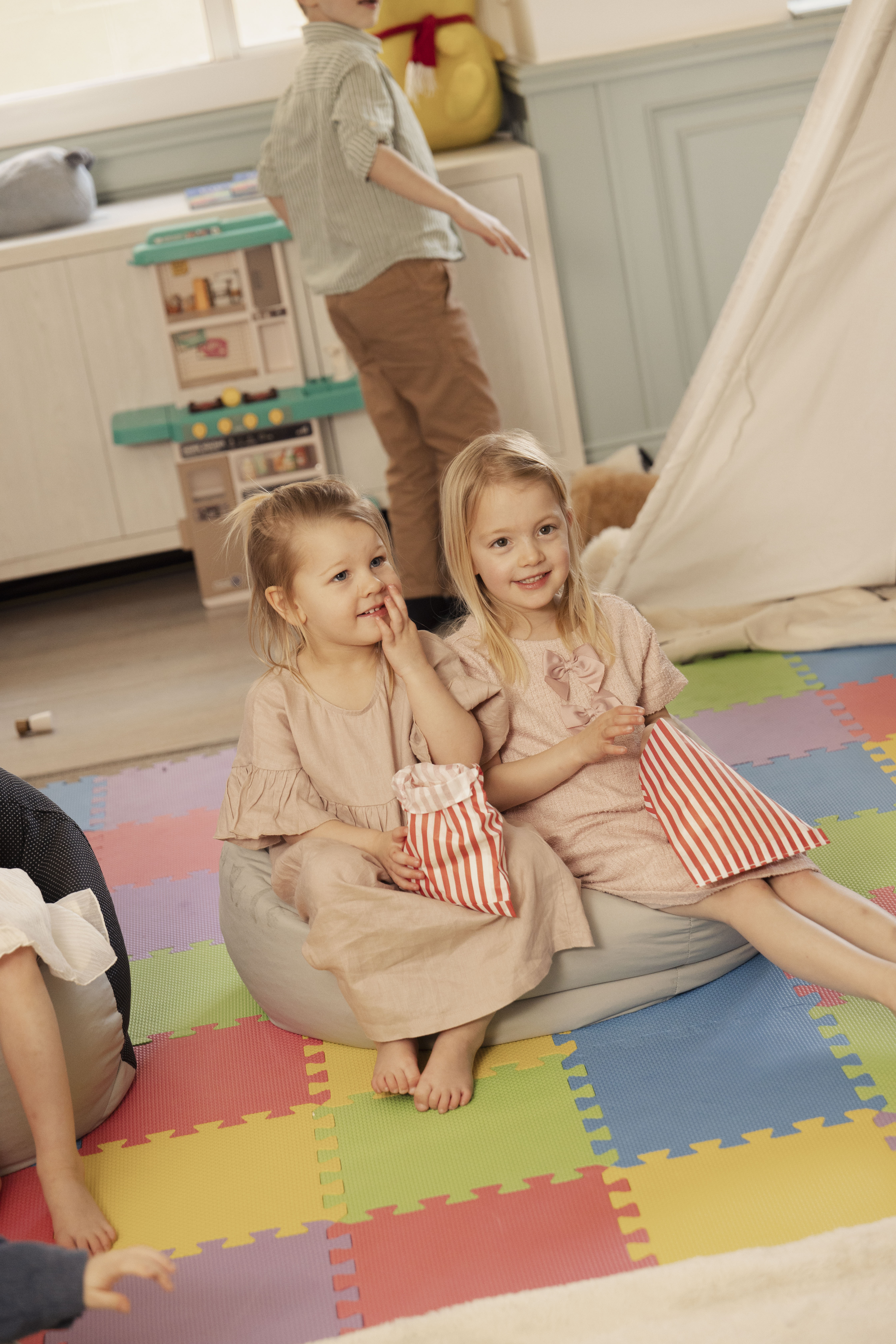 Two blonde girls on beanbags holding striped bags in a colorful playroom.