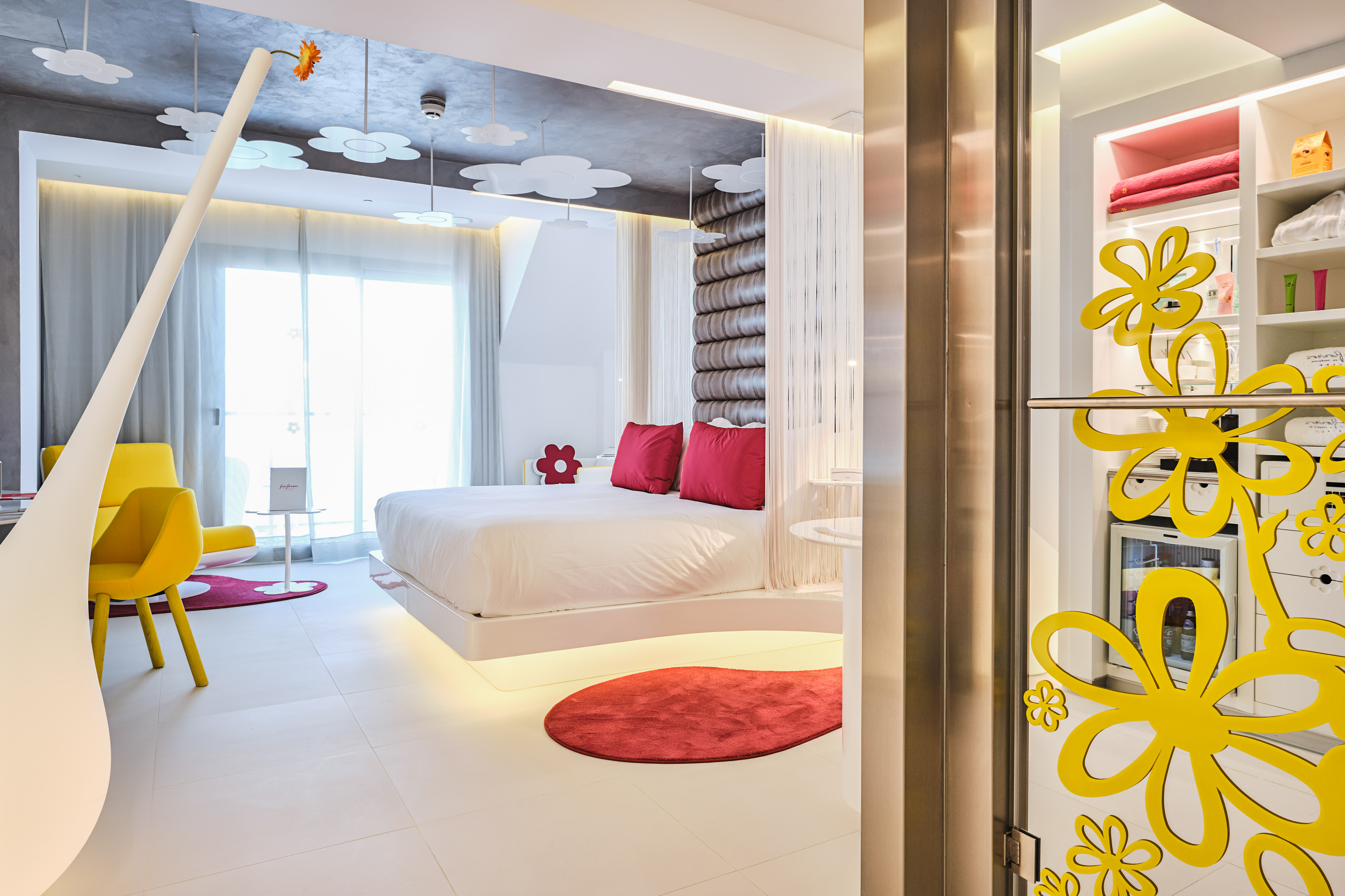 Modern hotel room with white bed, red accents, yellow chairs, floral decor.