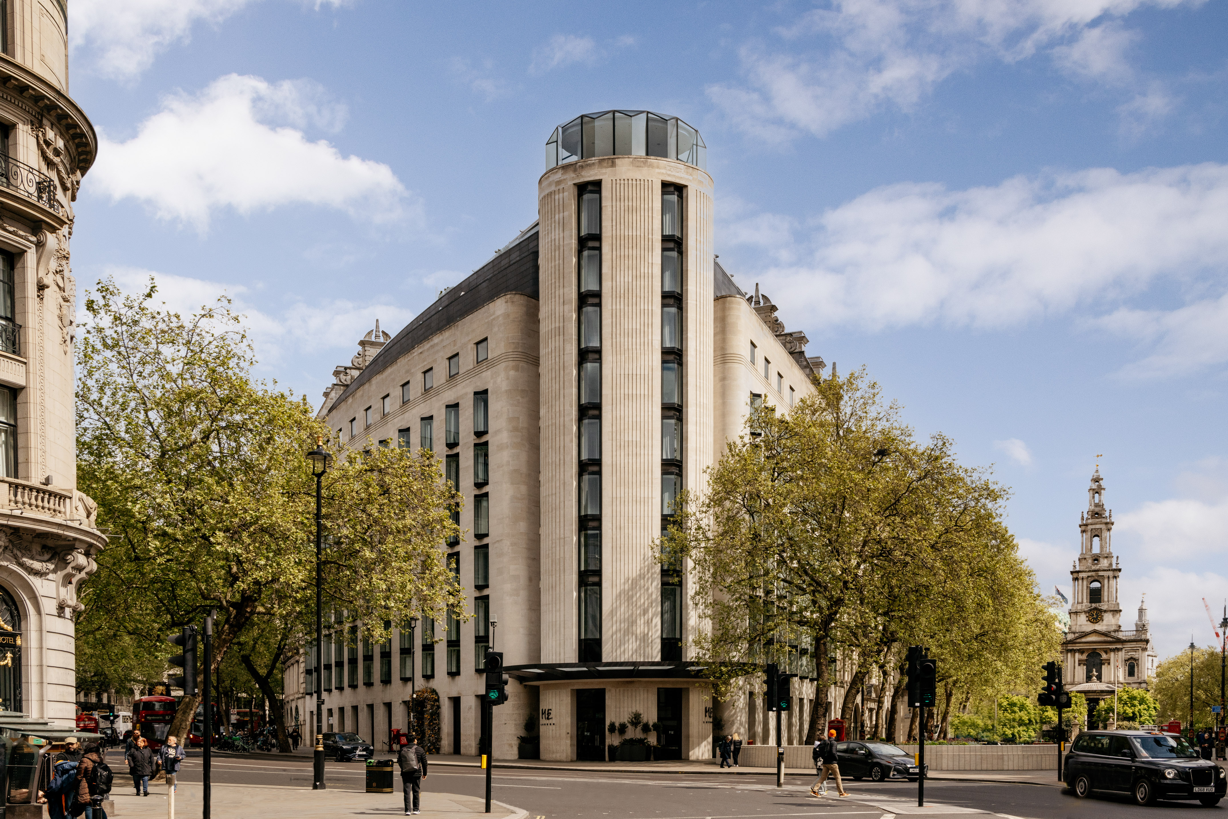 Modern ME London Hotel with a glass tower, street, and trees on a sunny day.