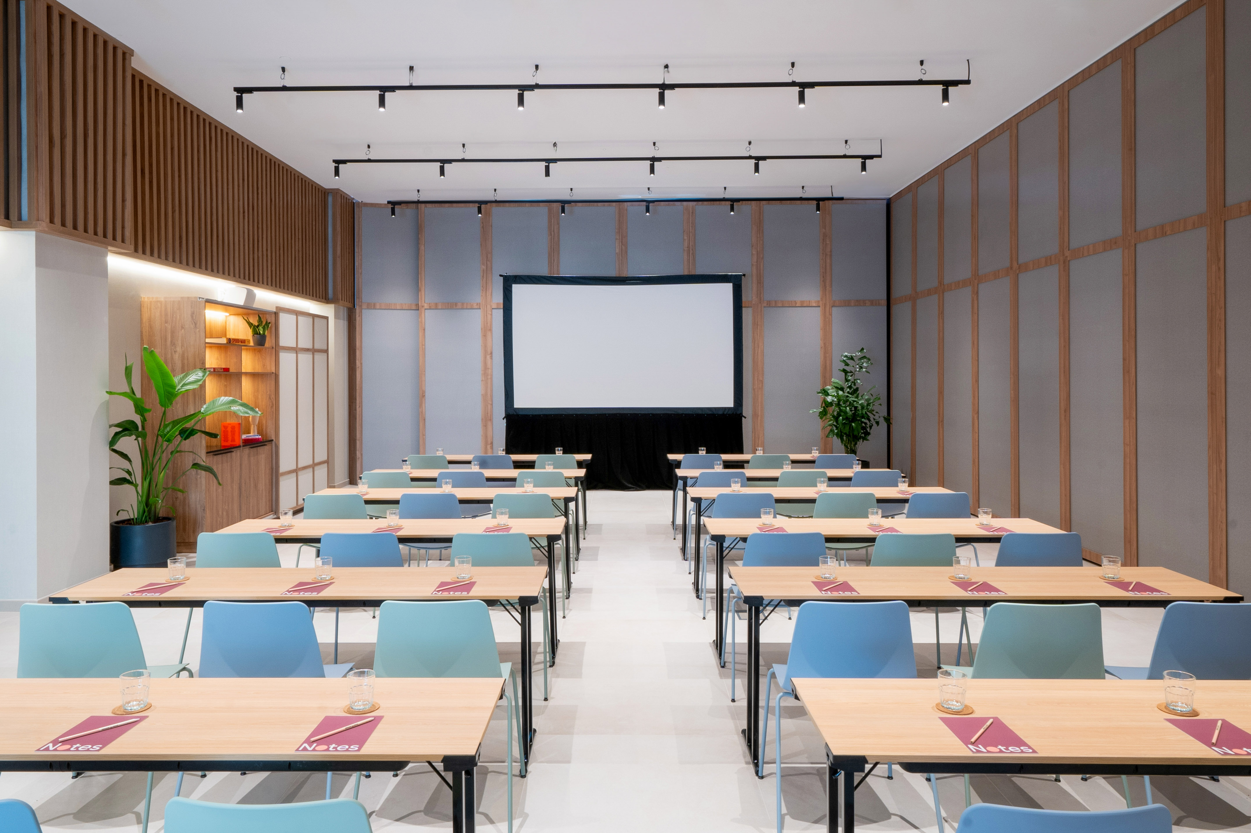 Modern hotel meeting room with classroom setup, light wood tables, projection screen.