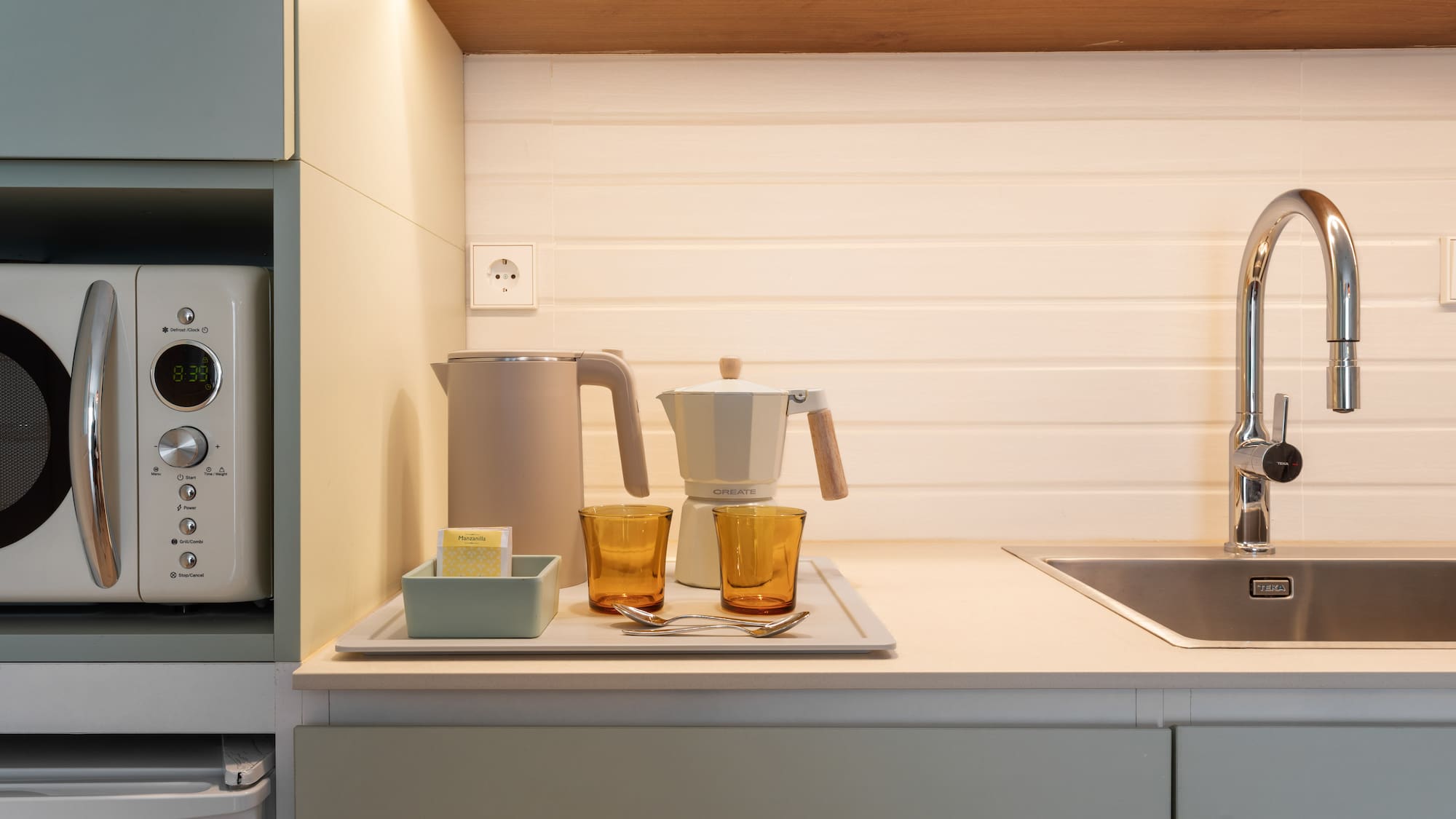 Modern kitchen counter with microwave, kettle, moka pot, sink, and glasses.