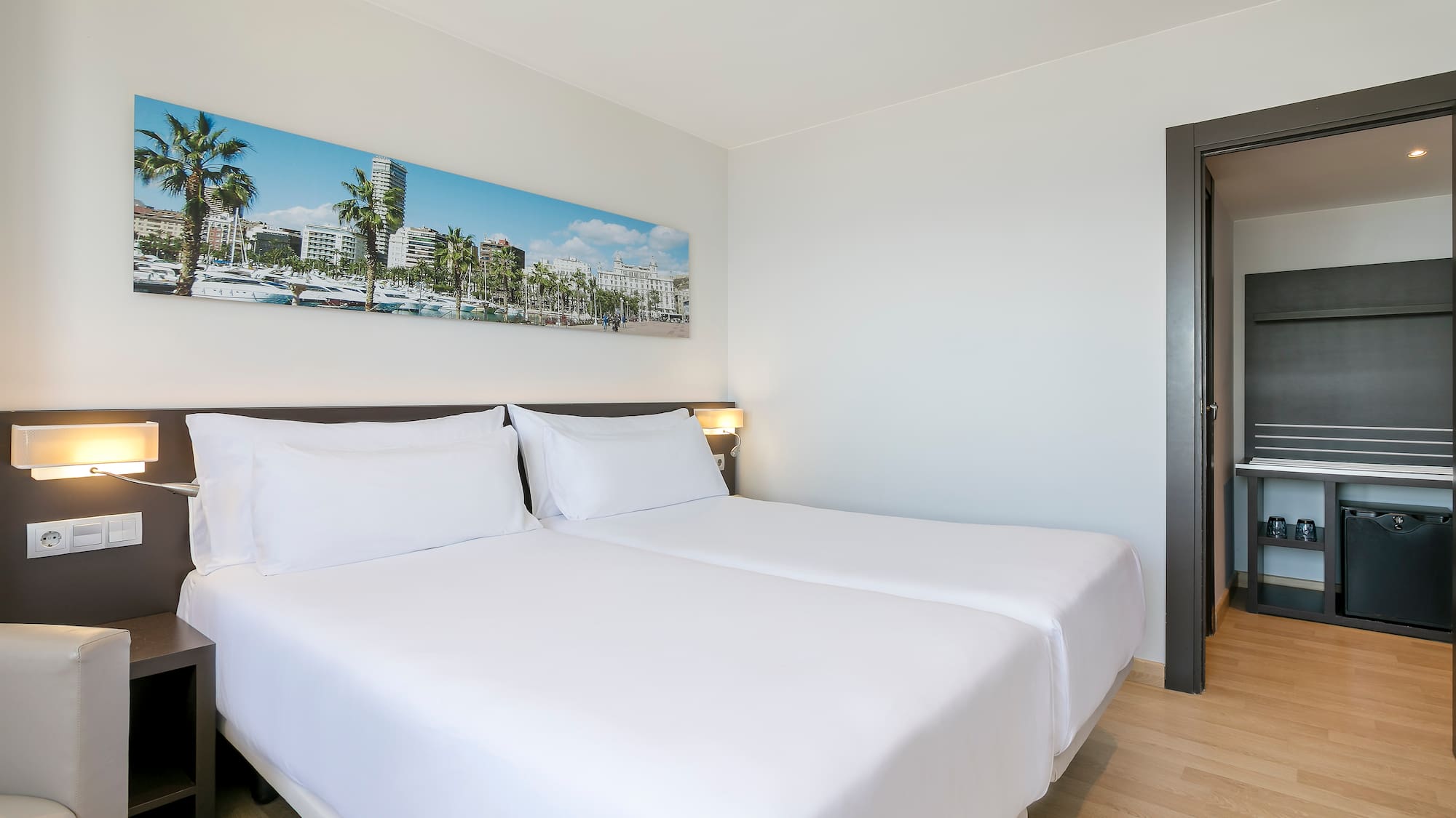 Modern hotel room with twin beds, white bedding, cityscape art, mini-bar.