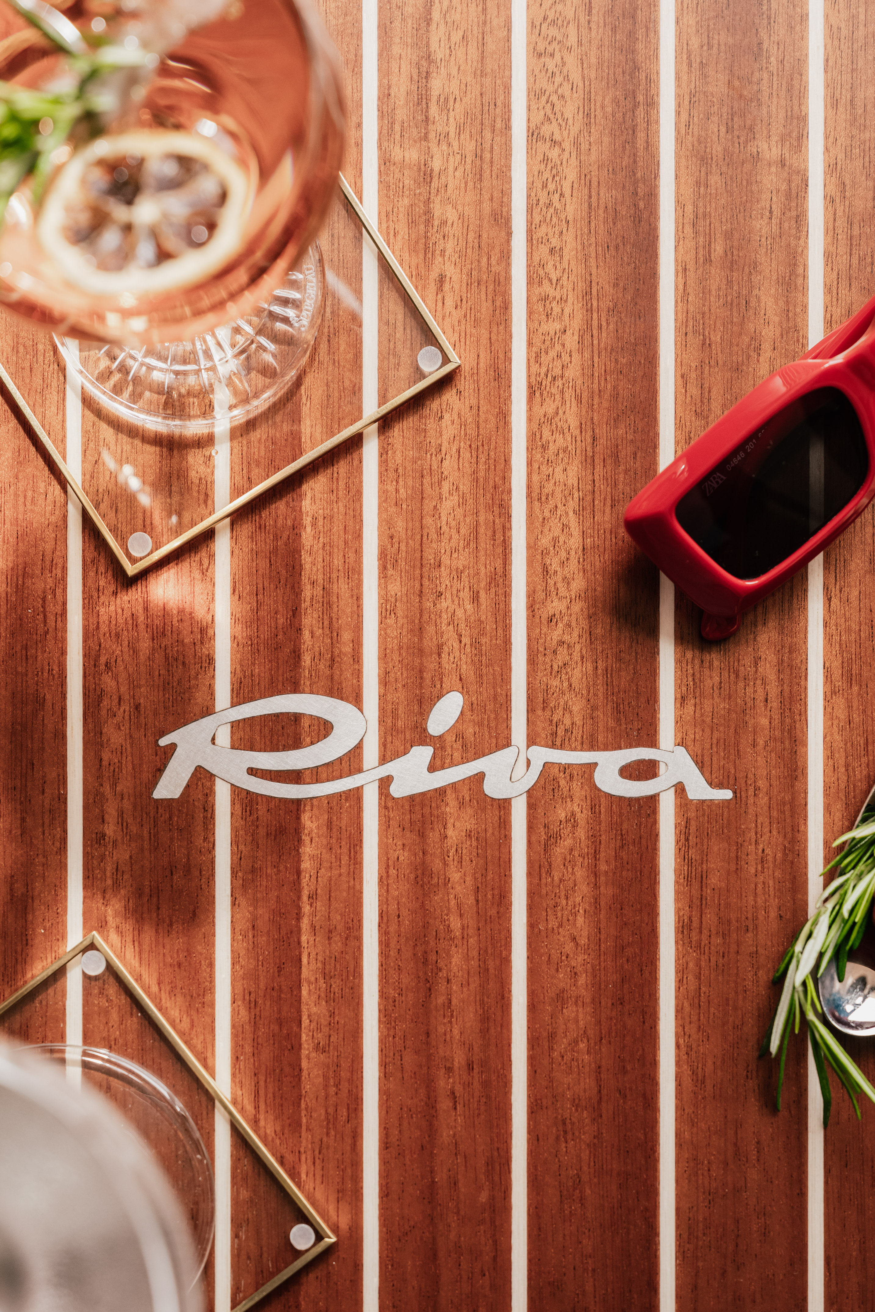 Riva logo on teak yacht deck with cocktail, red sunglasses, and rosemary.