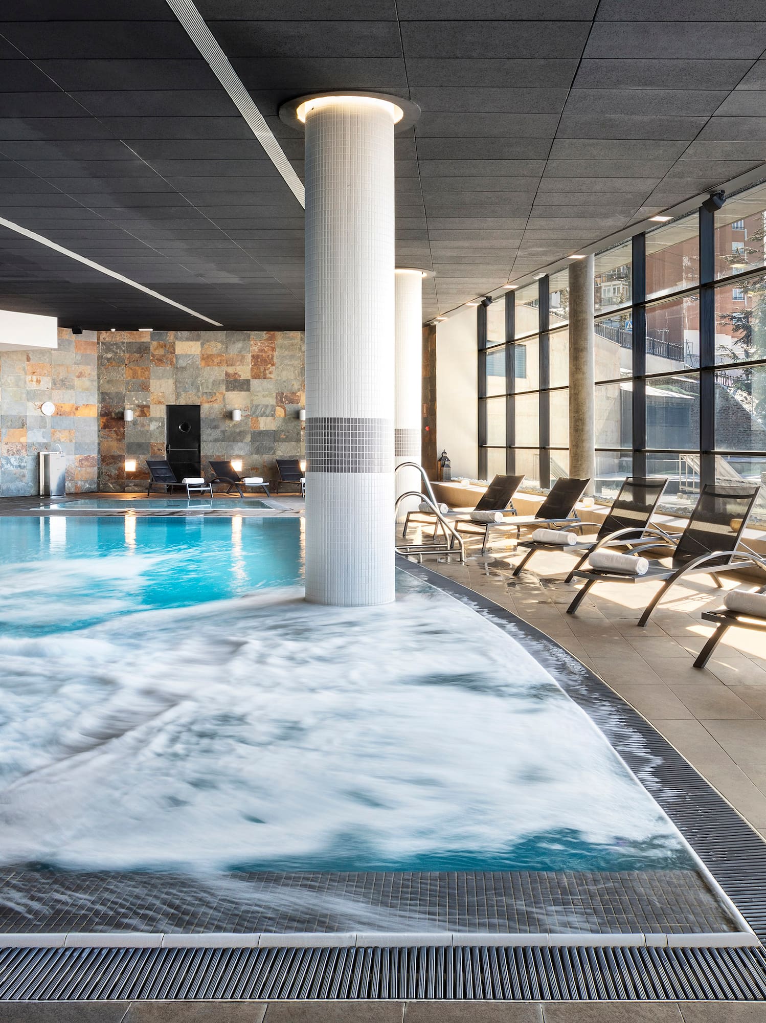 a pool with a waterfall and chairs in a room with windows