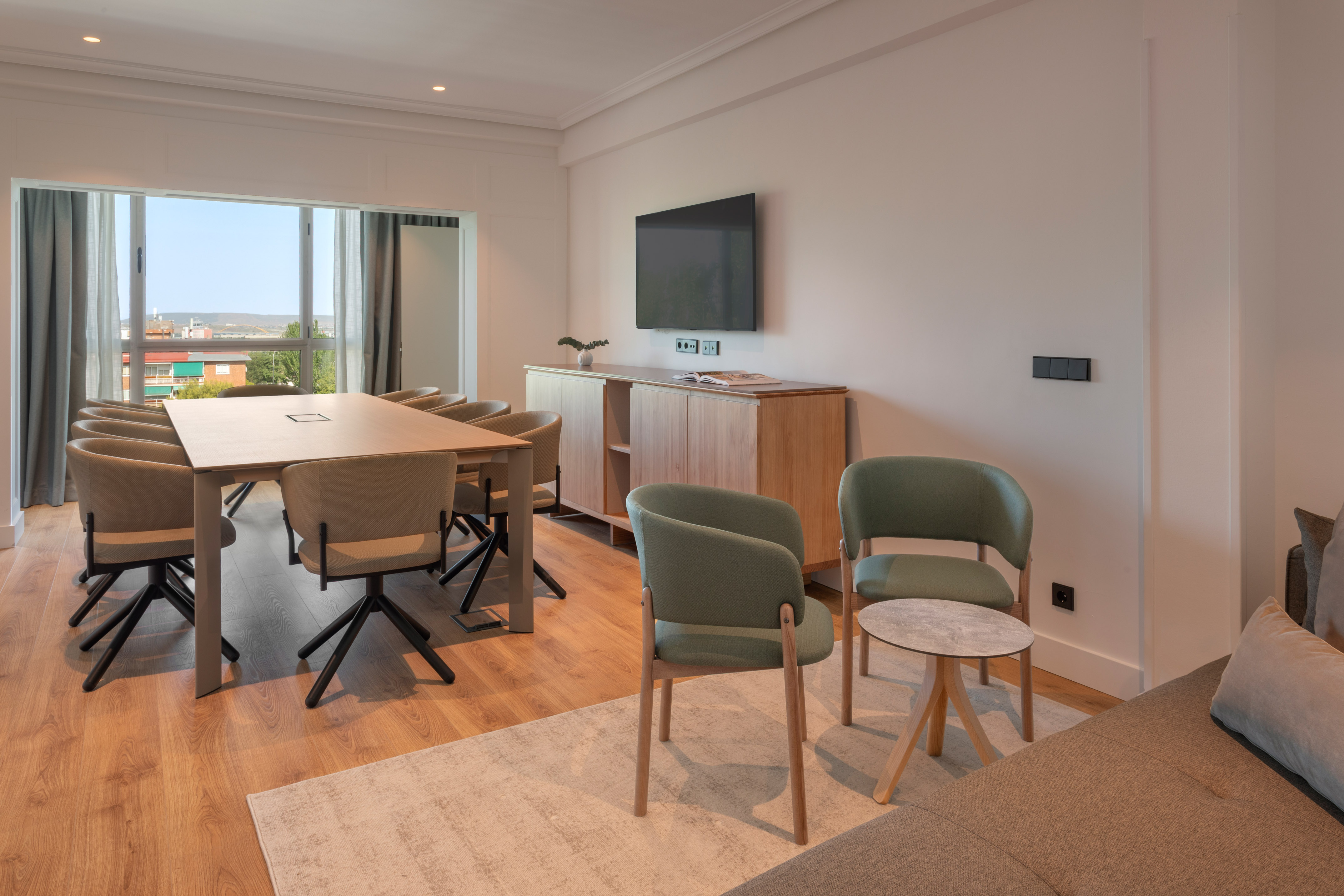 Modern hotel suite with long conference table, green armchairs, TV, and city view.