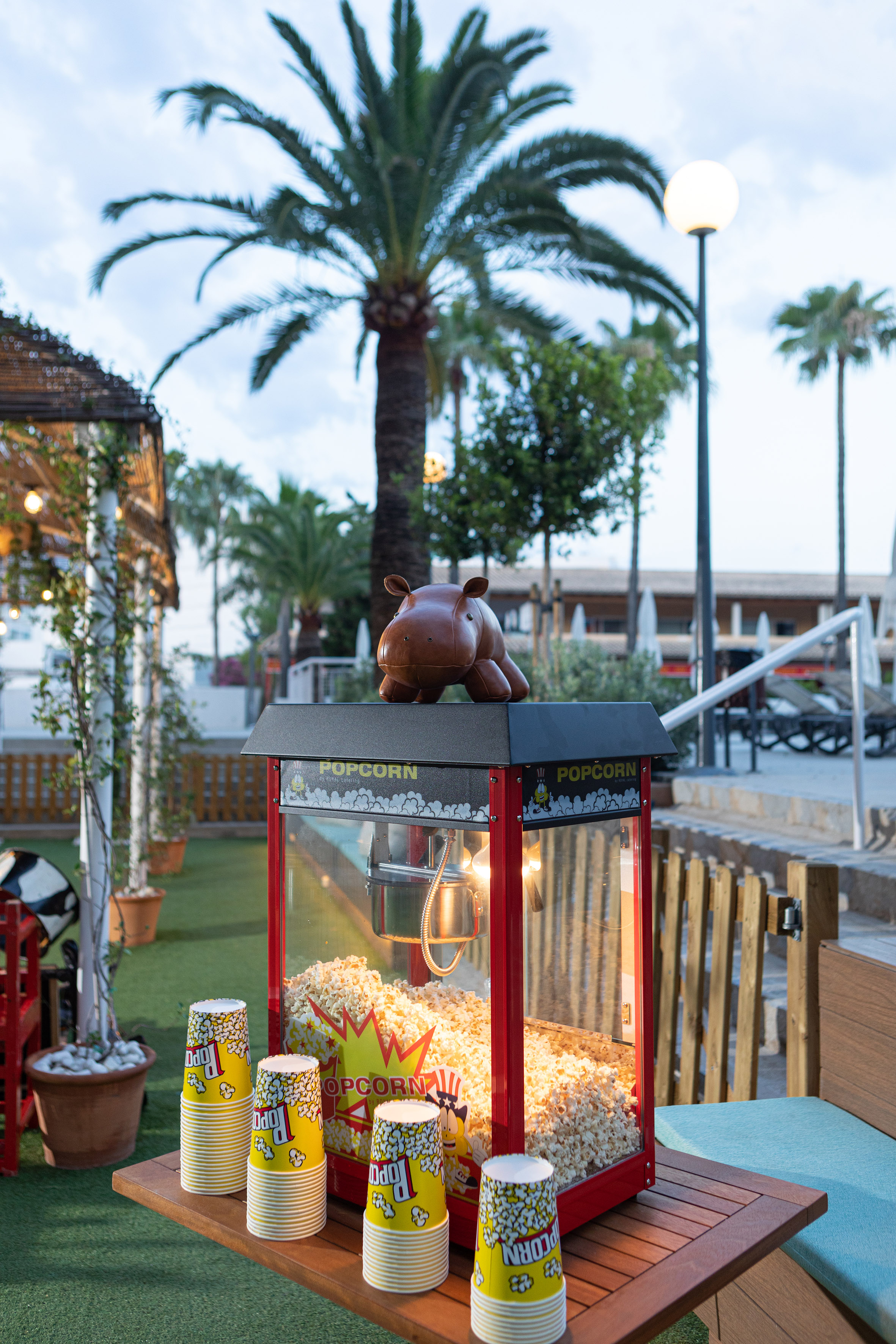 Red popcorn machine with popcorn, yellow cups, and a hippo toy at an outdoor resort.