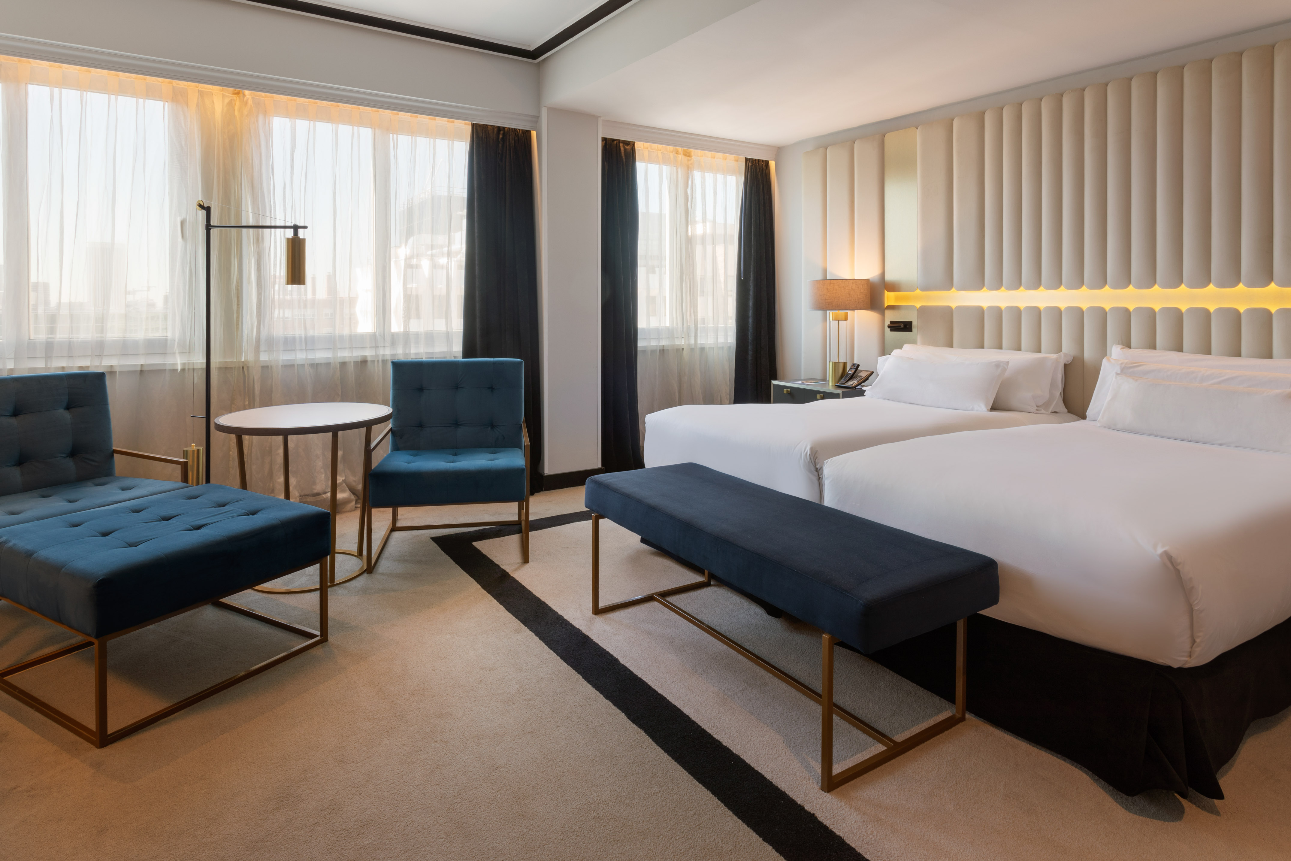Modern hotel room with twin beds, teal accents, and large window.