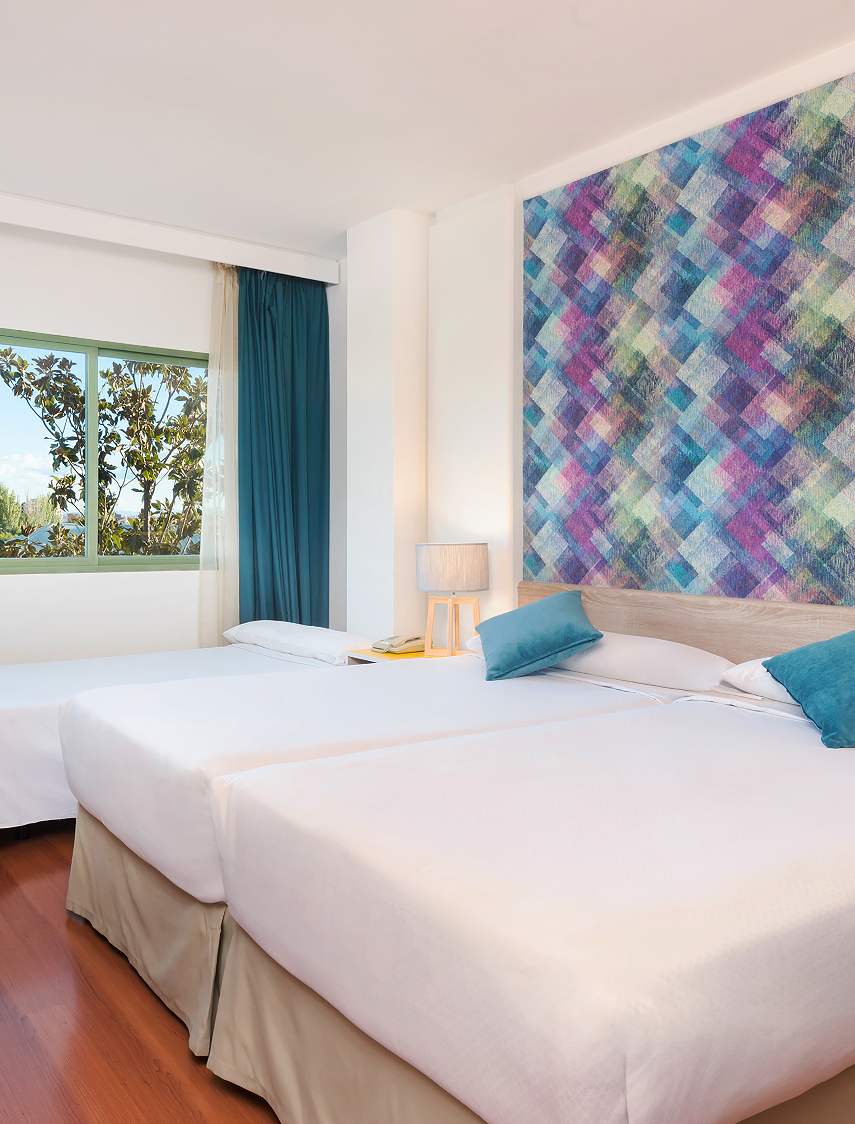 Modern hotel room with twin beds, geometric wall art, blue pillows, and window.
