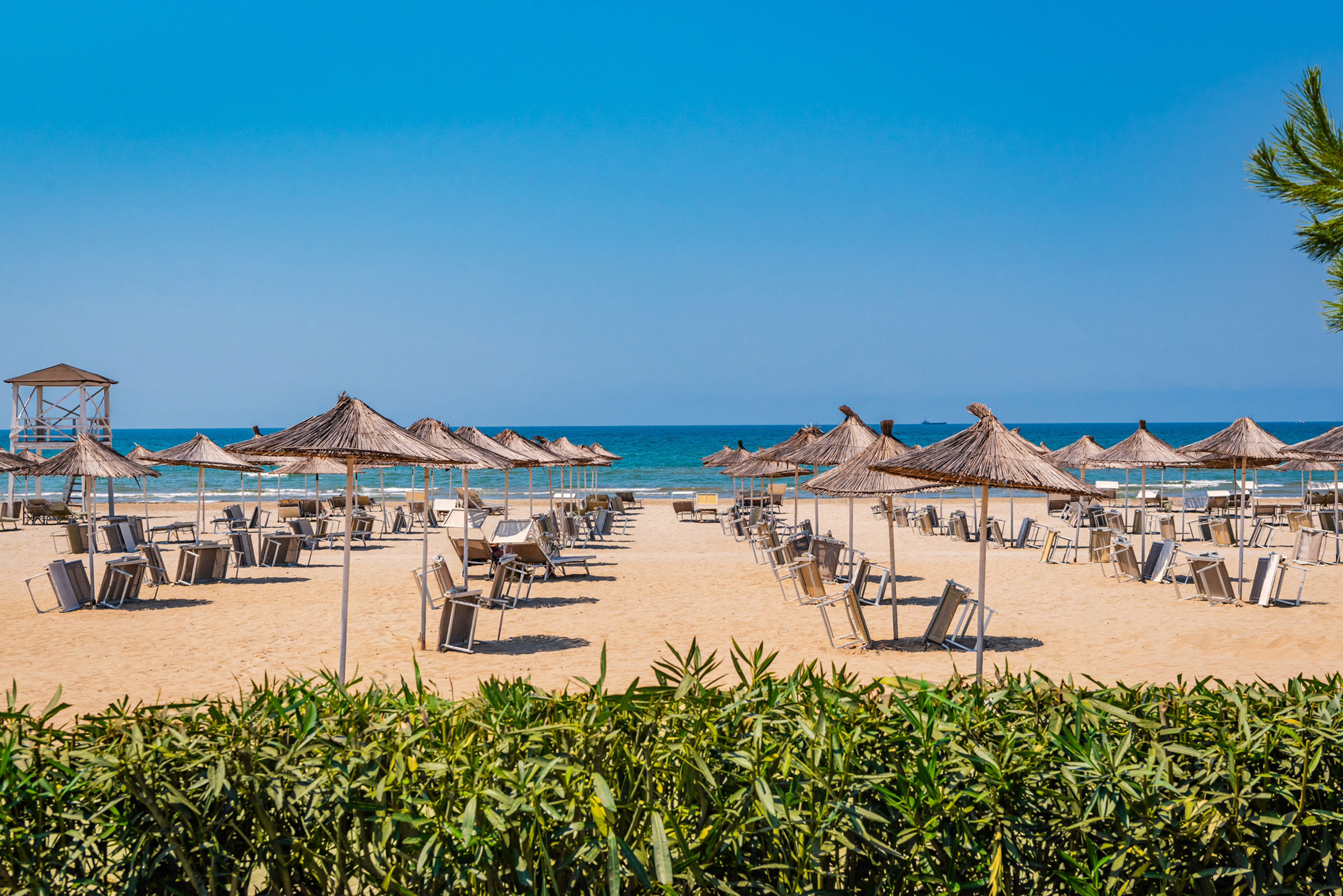 Sunny sandy beach with straw umbrellas, sunbeds, blue ocean, and lifeguard stand.