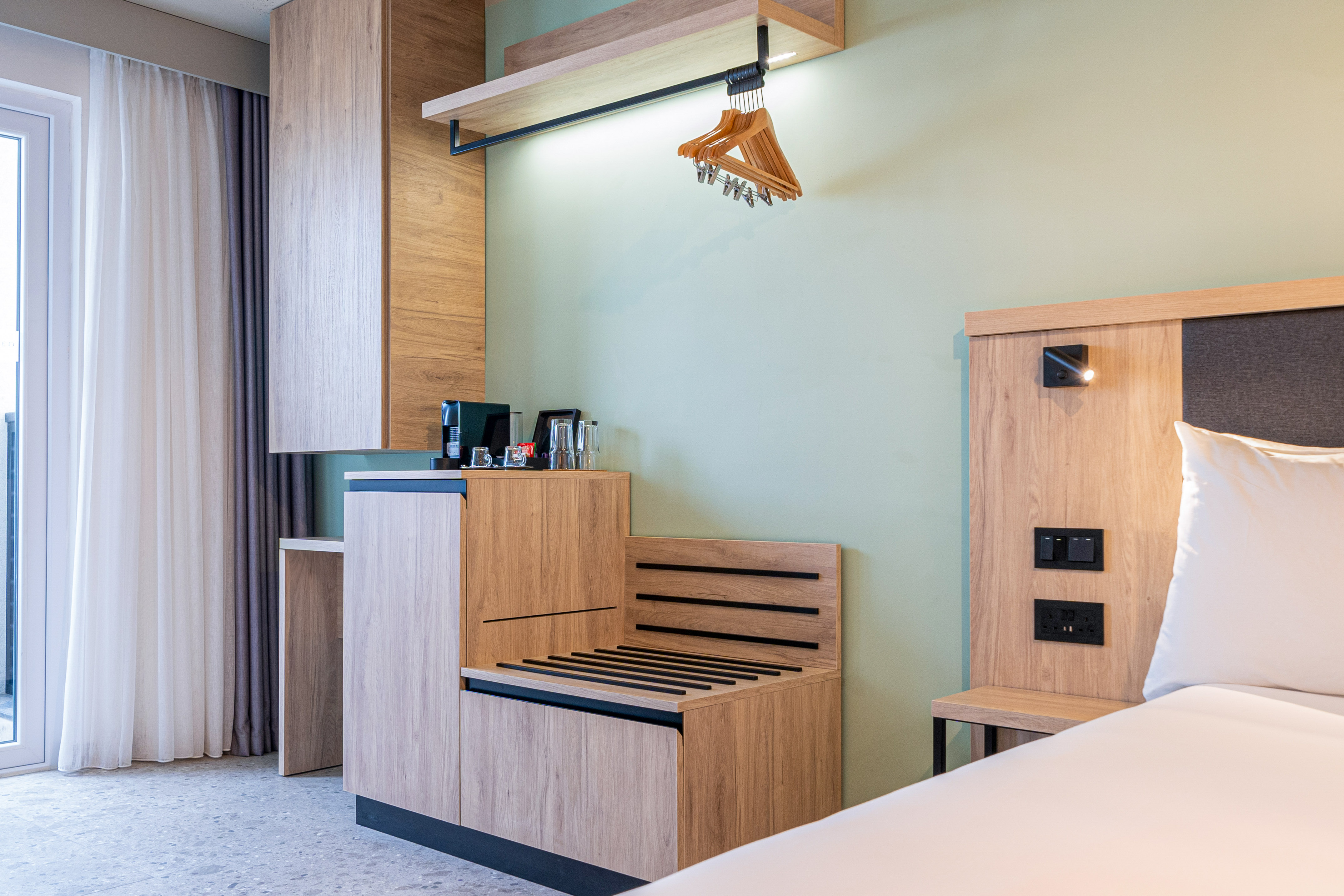 Hotel room with modern wooden furniture, coffee maker, luggage rack, green wall.