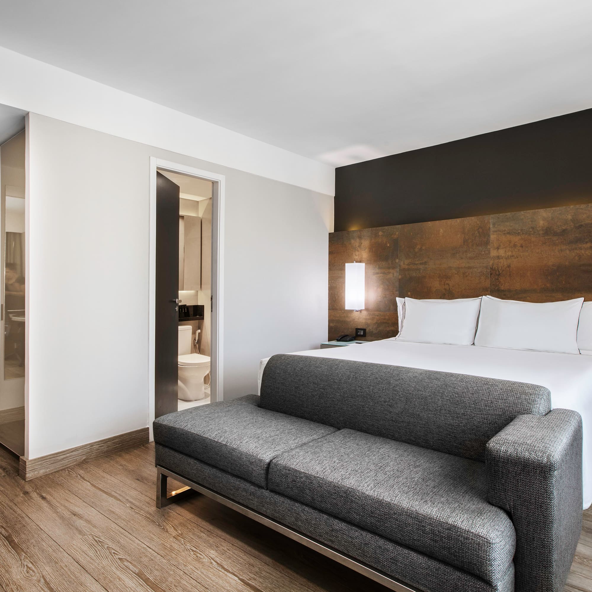 Modern hotel suite with bed, grey sofa, wood floor, and bathroom.