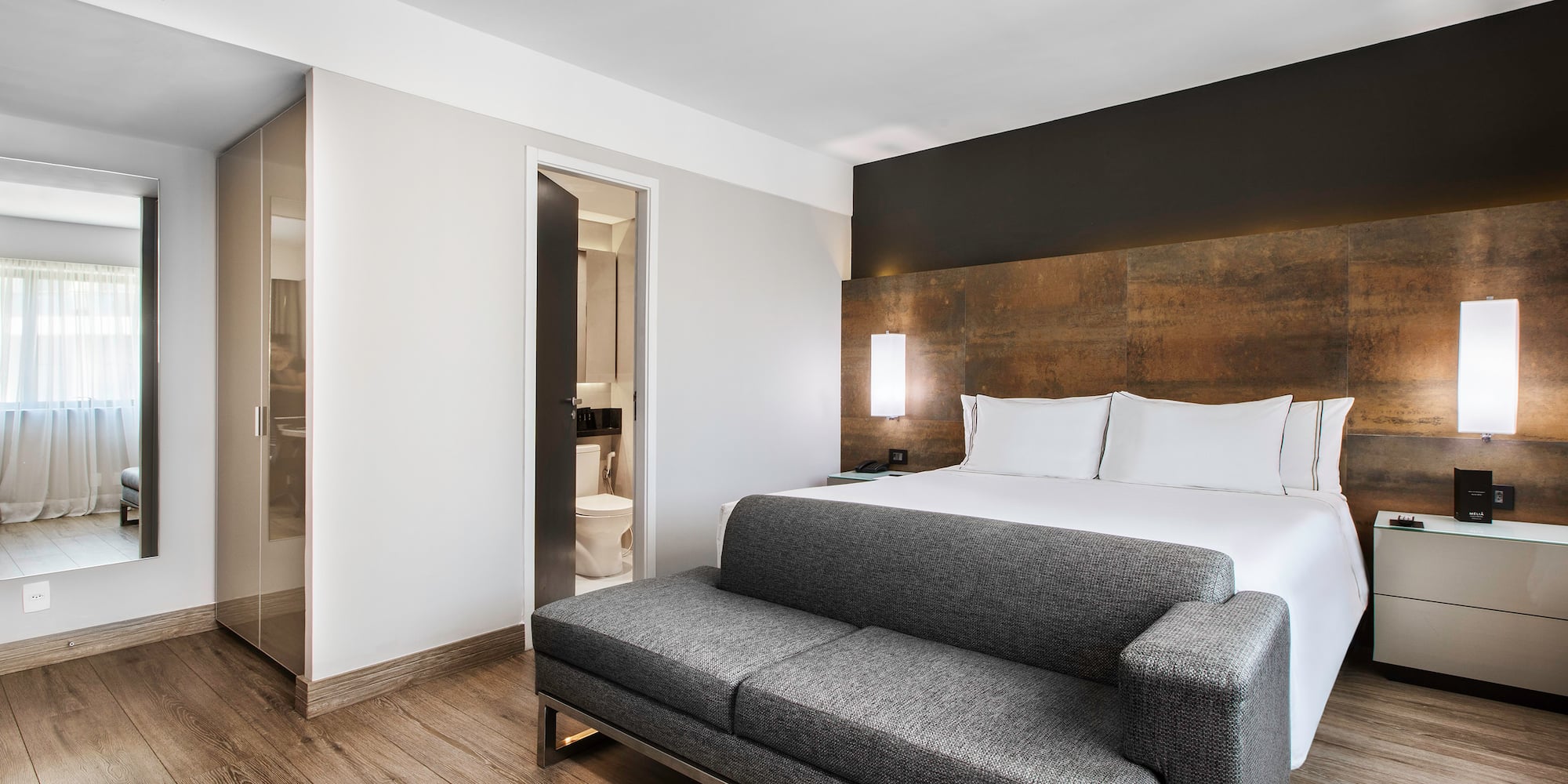 Modern hotel bedroom with large bed, gray sofa, wood floor, and ensuite bathroom.