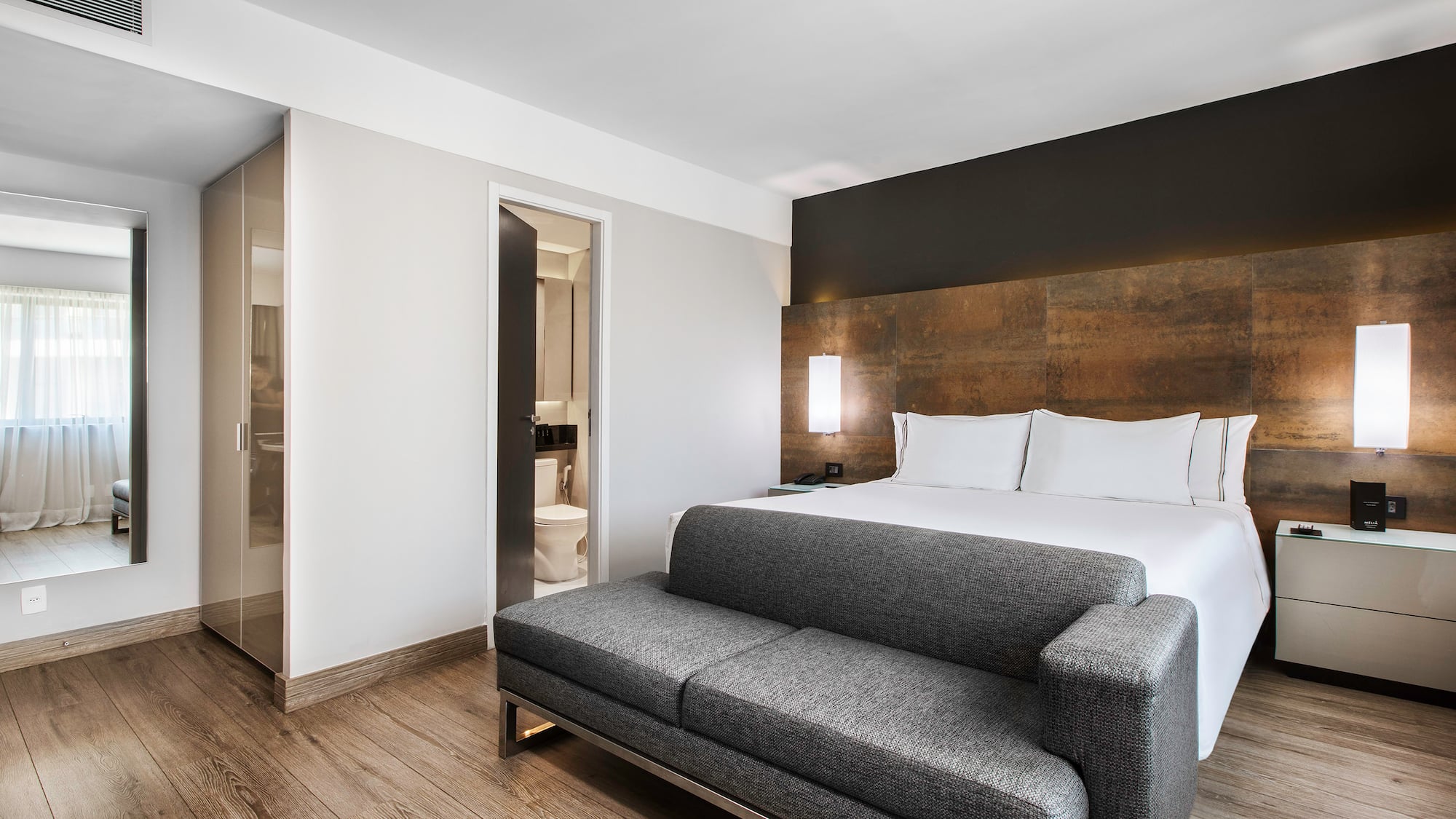 Modern hotel suite with bed, grey sofa, wood floor, and bathroom.
