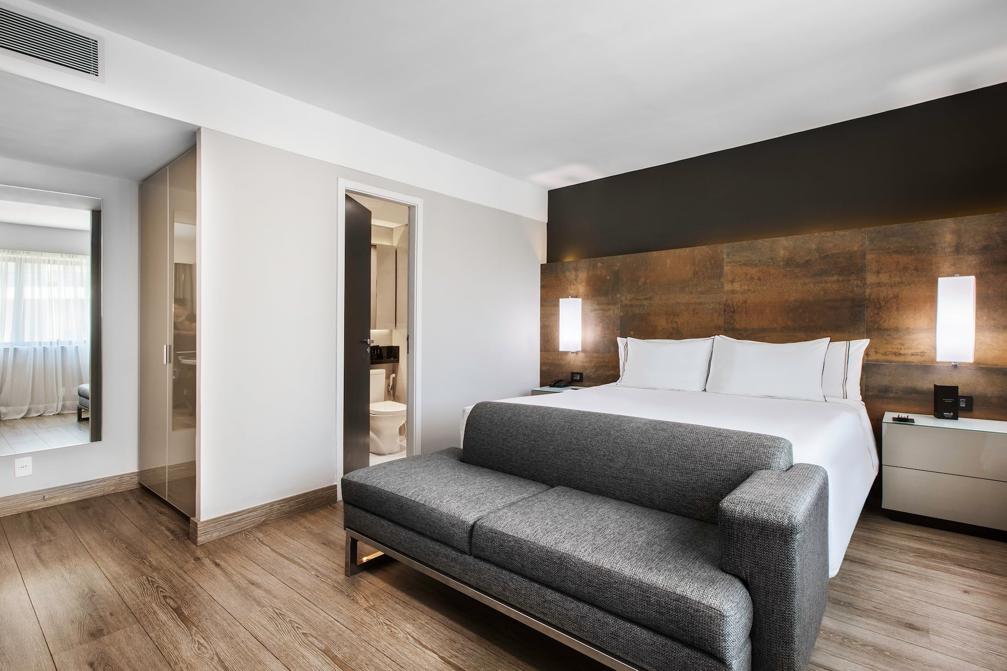 Modern hotel bedroom with white bed, gray sofa, wood floor, and open bathroom.