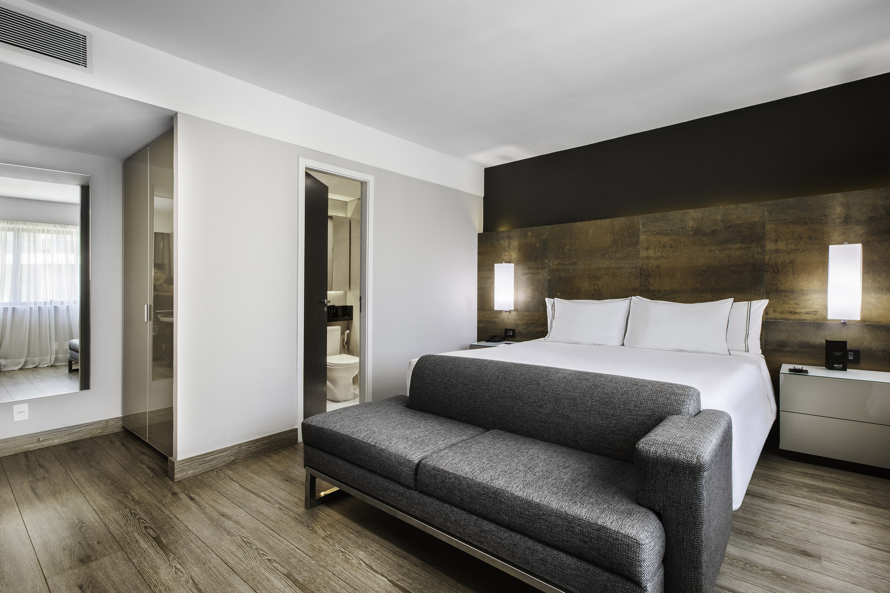 Modern hotel bedroom with white bed, gray sofa, wood floor, and open bathroom.