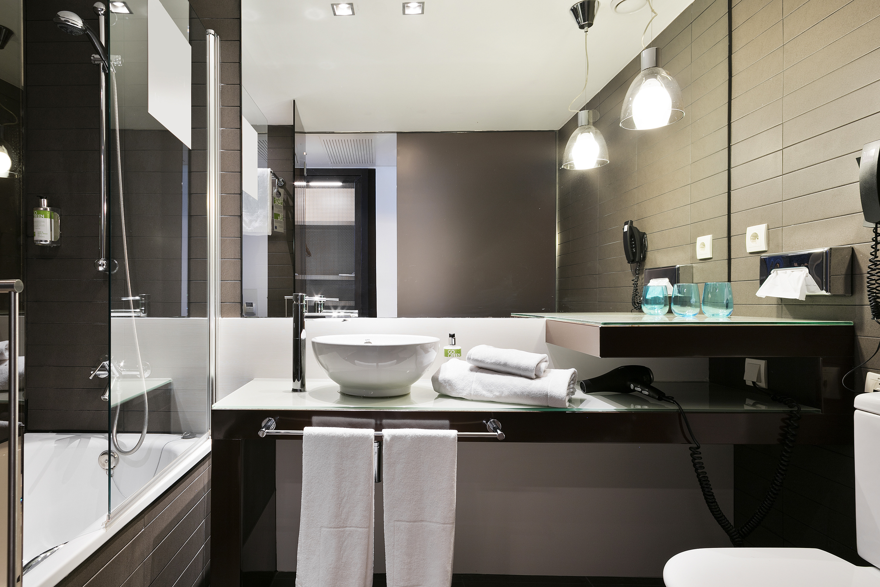 Modern hotel bathroom with bathtub, vessel sink, vanity, and brown tiled walls.
