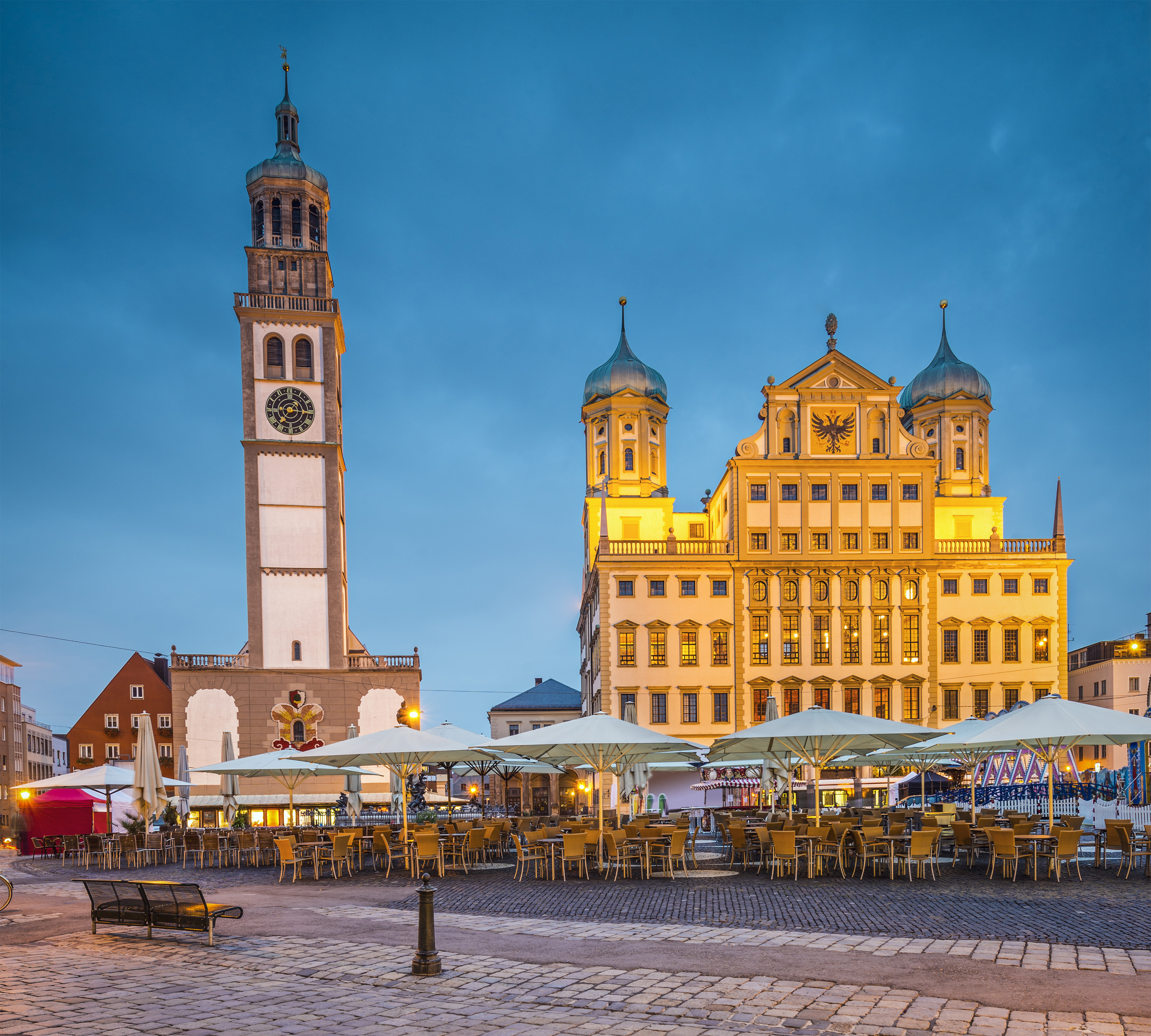 Augsburg Rathaus, Perlachturm and outdoor cafes on cobblestone square at dusk.