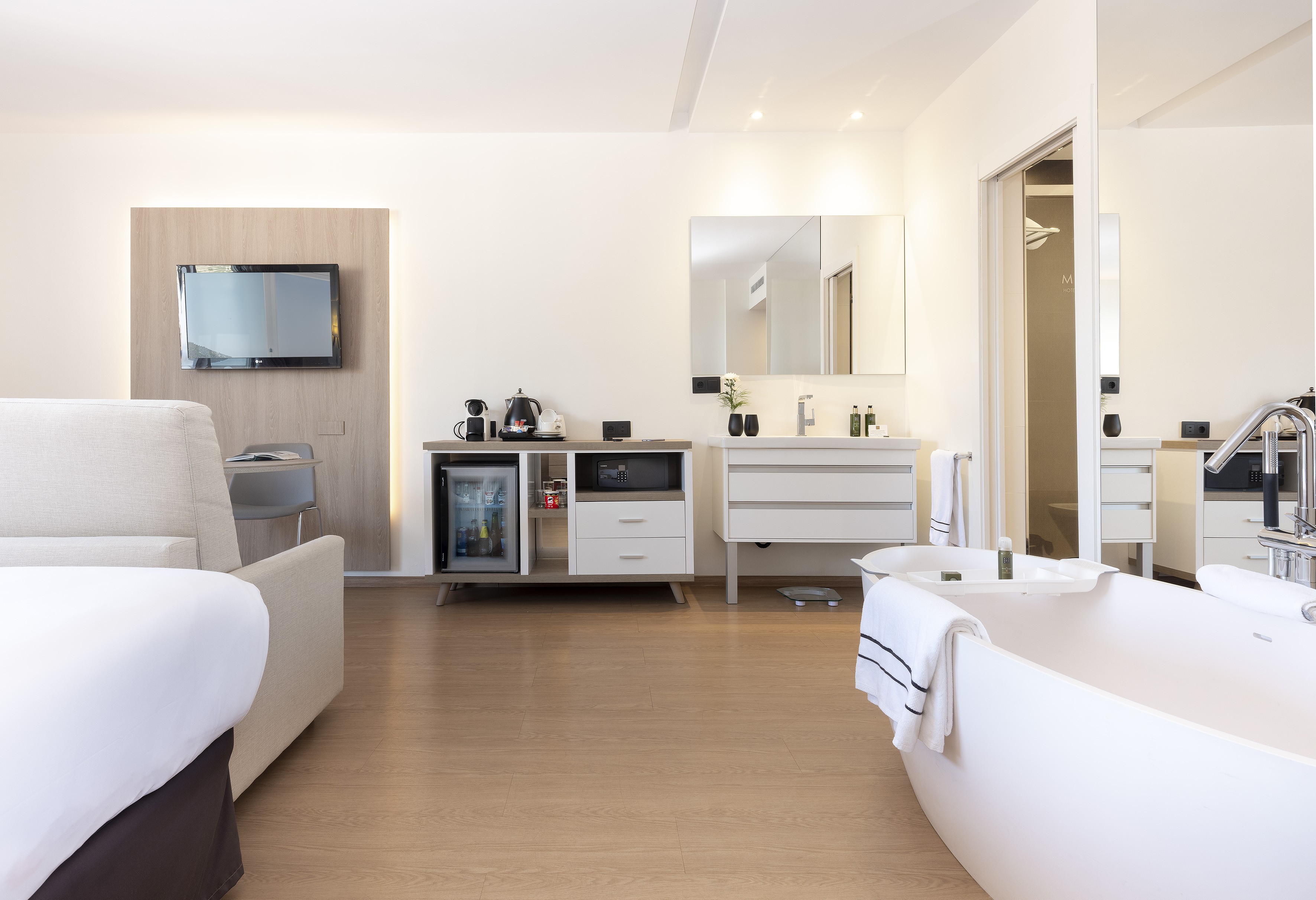 Modern hotel suite with bed, freestanding tub, TV, minibar, bathroom vanity.
