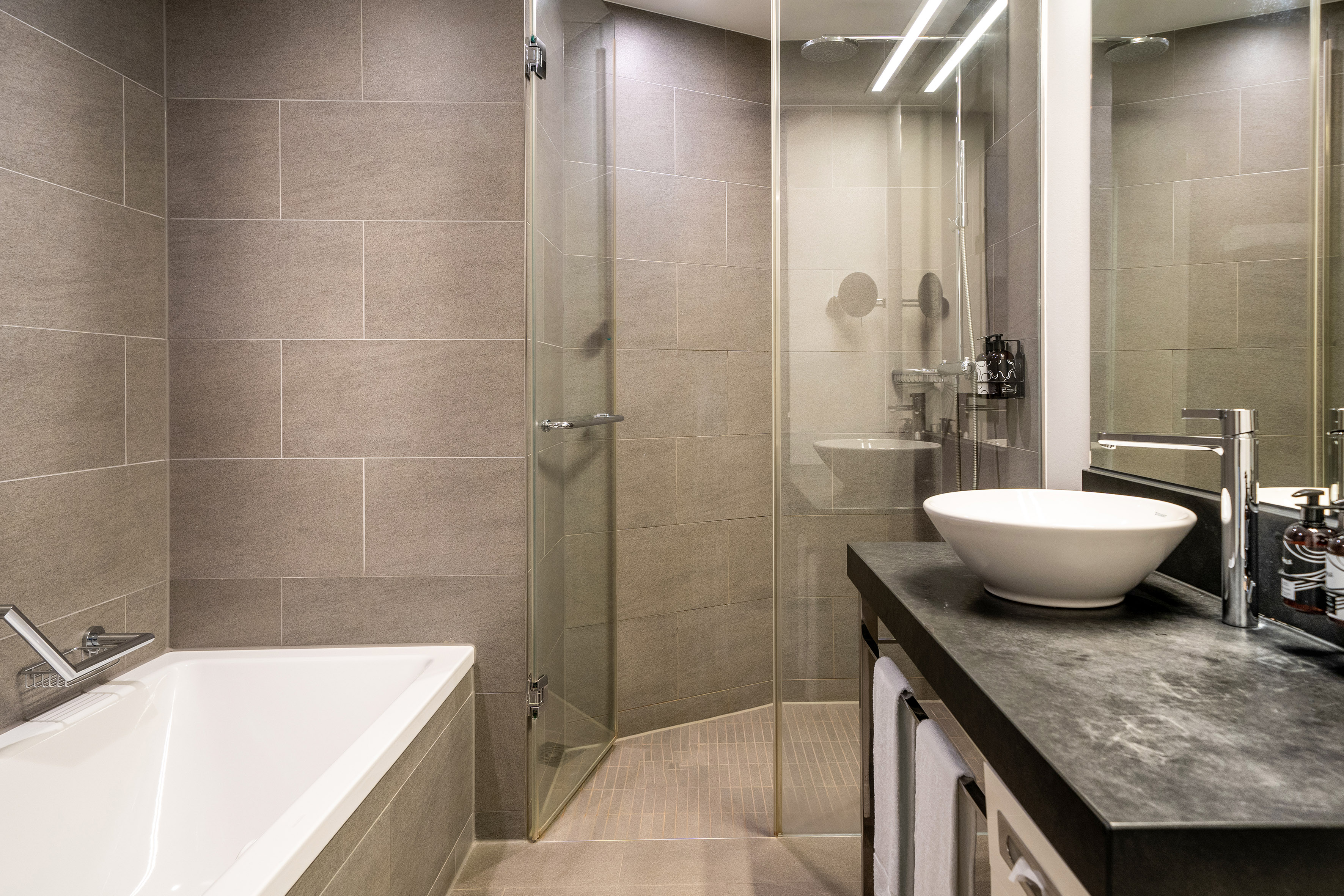 Modern hotel bathroom with gray tile walls, bathtub, glass shower, and vanity.