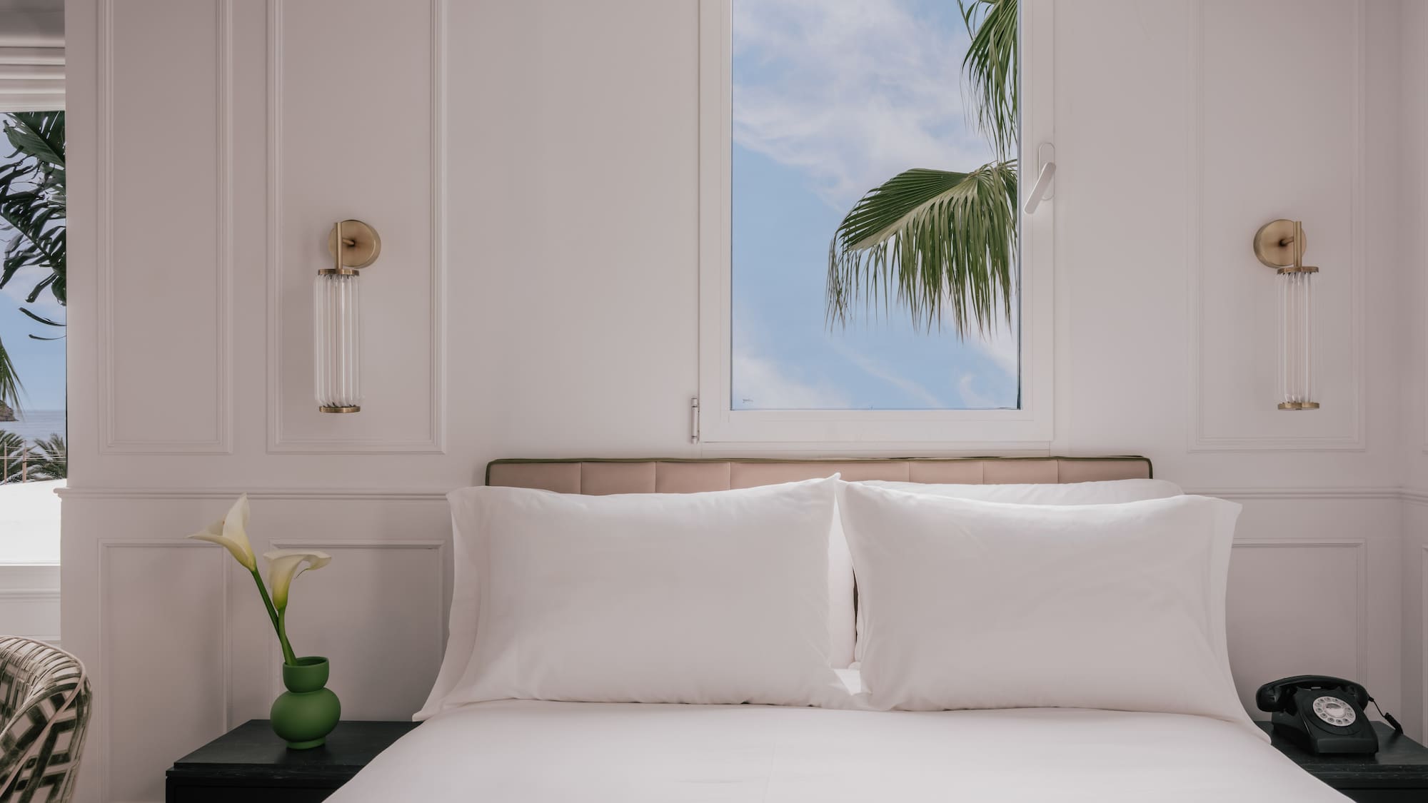 Modern white bedroom with clean bed and tropical palm tree window view.